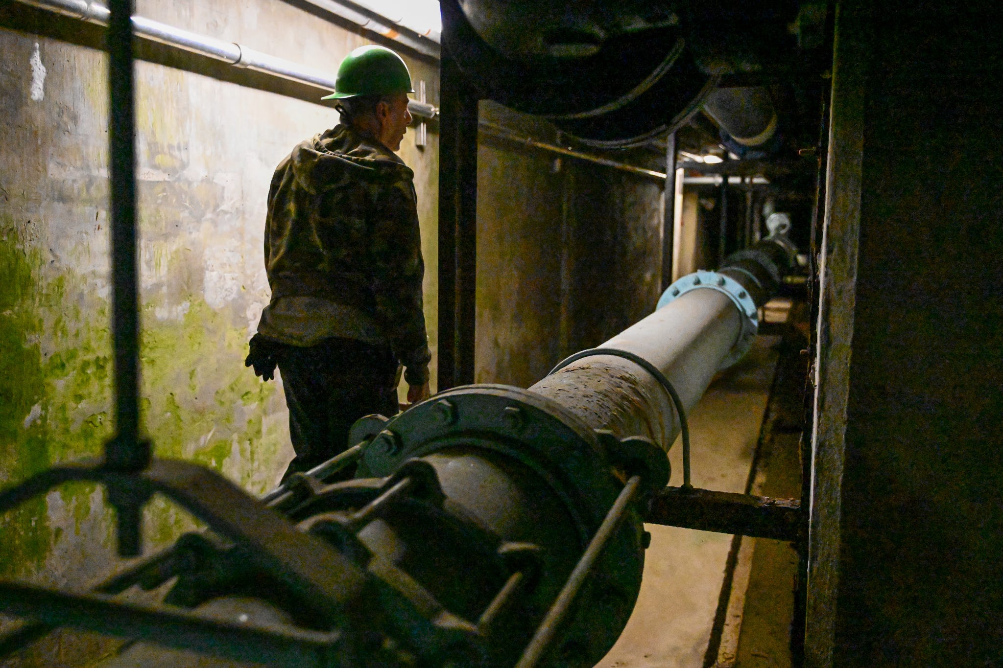 a man in a camouflaged hoodie and green hard hat walks in a gloomy tunnel under the glow of singular overhead LED lights. he is seen between brackets of a wall-mounted pipe filled with water as he walks forward into the darkness. pic by Nicole Harvel.