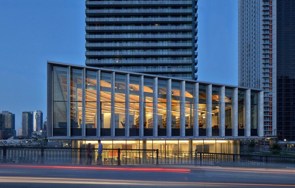 Photograph of a modern building lit from within at dusk. On a glassed-in one-story pedestal sits a trapezoidal form inside of which are rows of library shelves. A modern residential tower rises behind it. Photograph of a modern building lit from within at dusk. On a glassed-in one-story pedestal sits a trapezoidal form inside of which are rows of library shelves. A modern residential tower rises behind it.