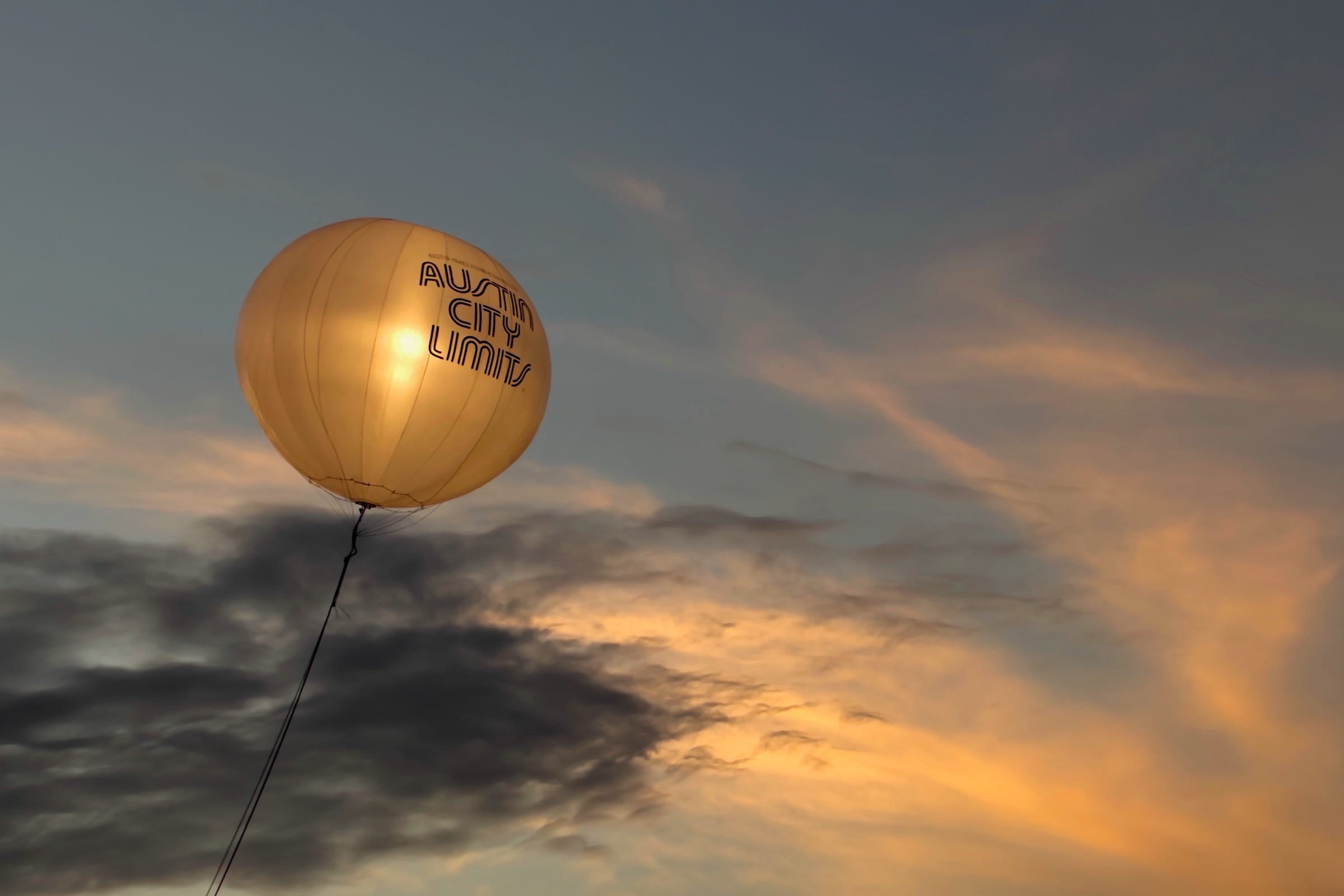 A lighted balloon attached to a wire with the words, Austin City Limits, floats against the sky with grey, and white clouds highlighted by the rays of the setting sun to the right of the photo