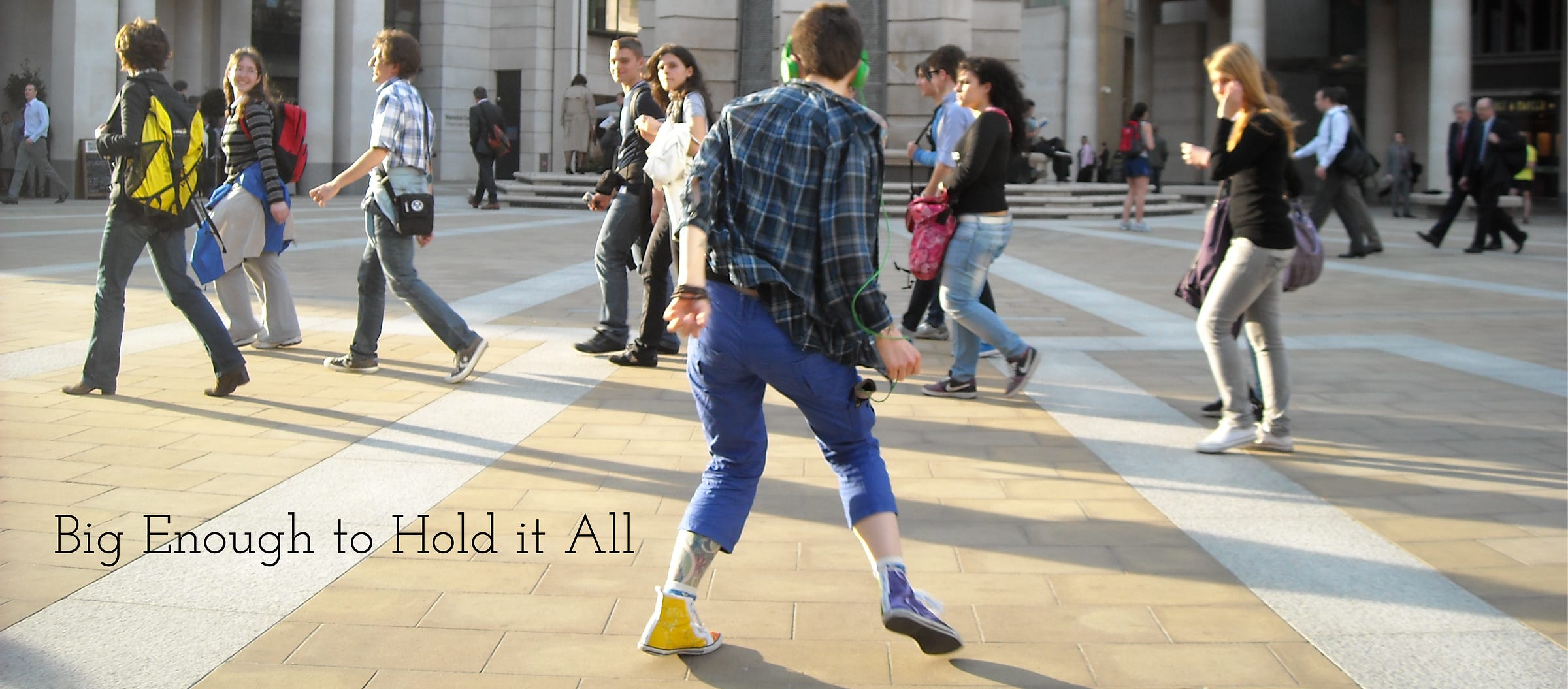 Photograph of the blog author dancing in a crowded public square. The author's back is to the camera. They are wearing a blue and black plaid shirt and blue capri pants. A tattoo shows on their left leg. On their feet are colourful hand painted canvas high tops. They have on bright green headphones. People walking past them in the background are glancing their way or looking forward in the direction they are walking. The title of the poem this image goes with, Big Enough to Hold it All, is in the bottom left corner of the image. 