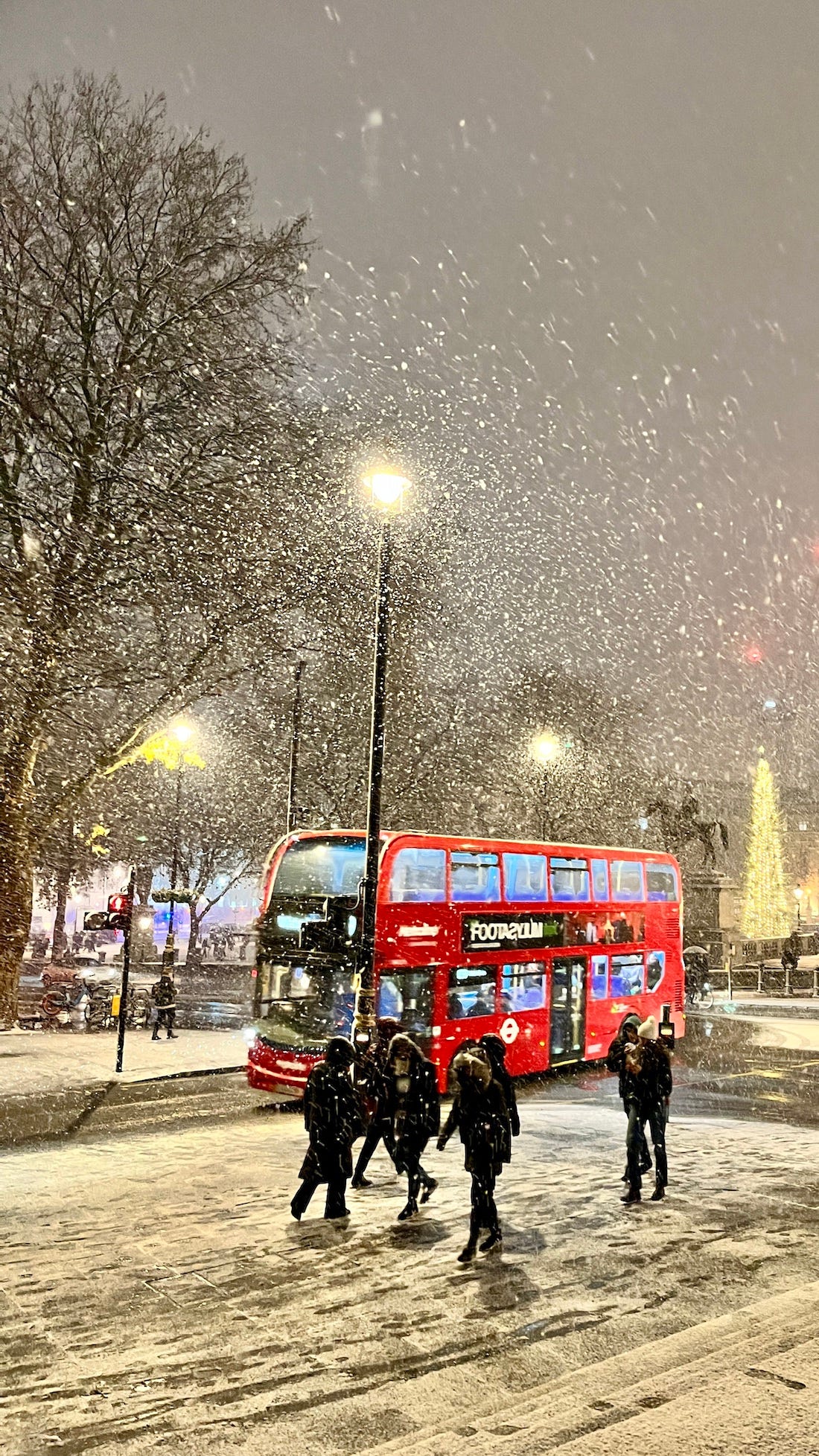 Image: a photo of the iconic red bus on the streets of London in Trafalgar Square, the street and building lights, and the Christmas tree, twinkling in the background, with snow falling in the foreground. 
