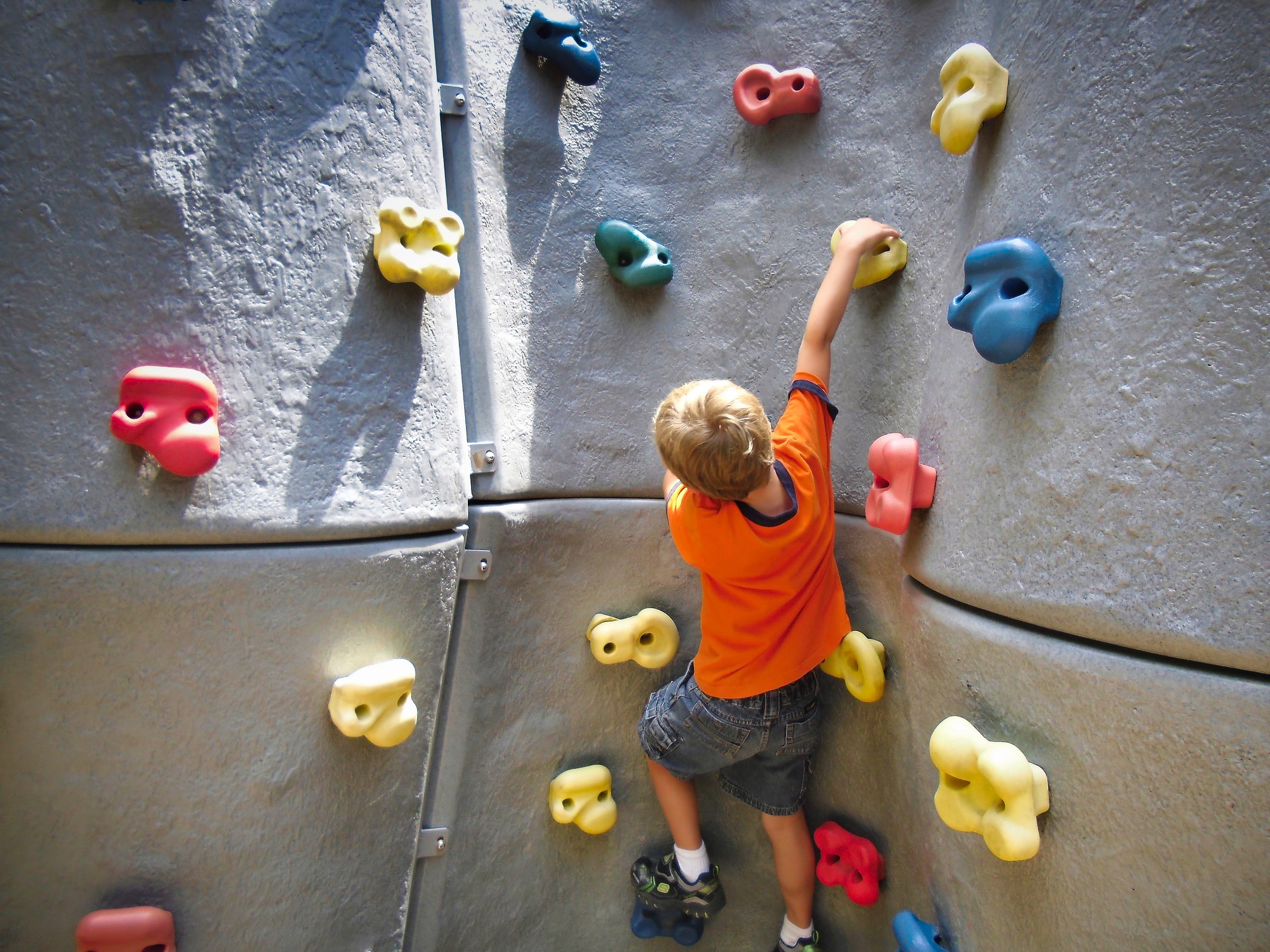 A young boy in an orange shirt and jean shorts reaching for the next grip on a rock climbing wall 
