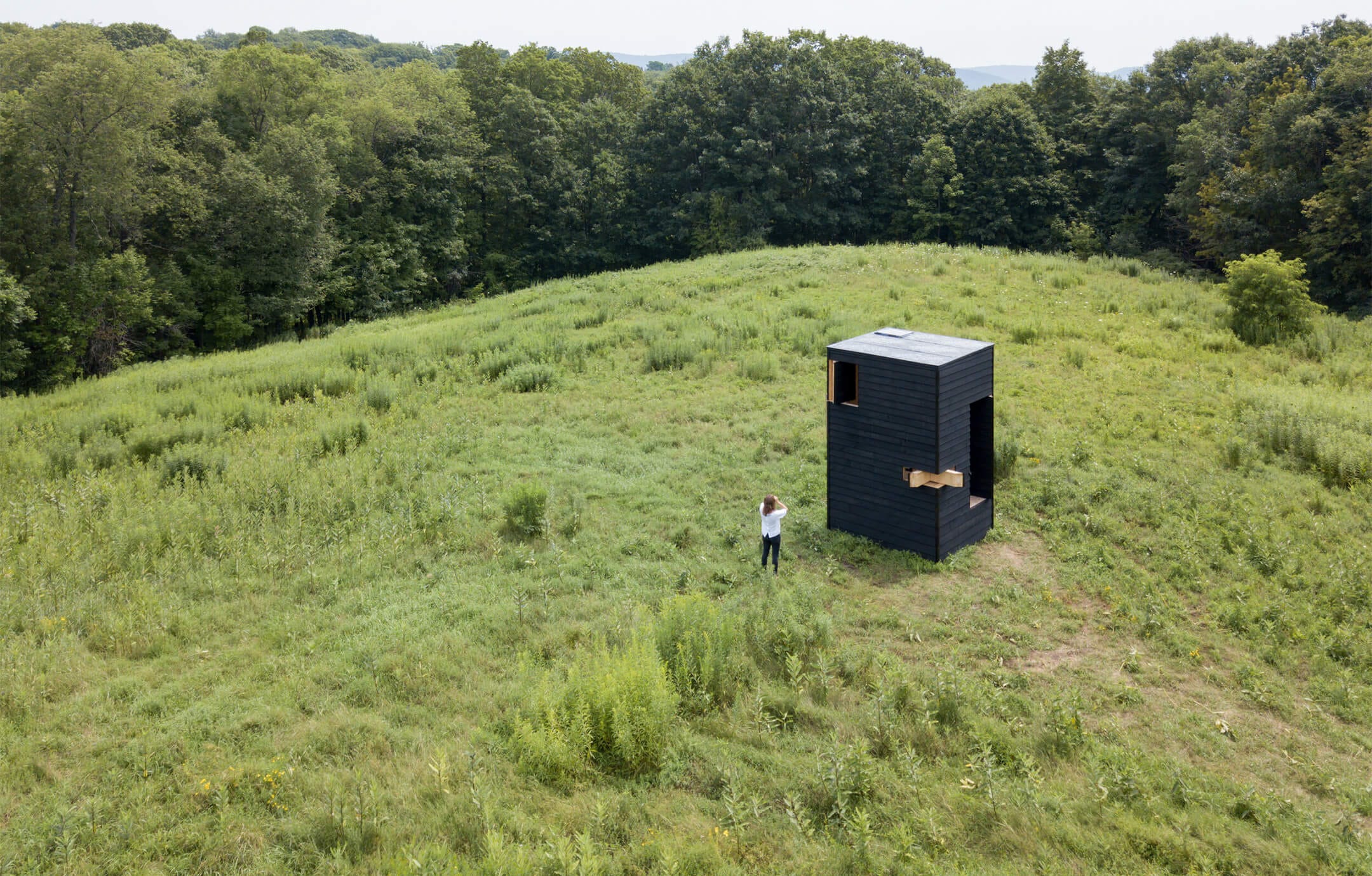 Drone-eye view of a cabin, in black-painted wood, in the middle of a grassy hill