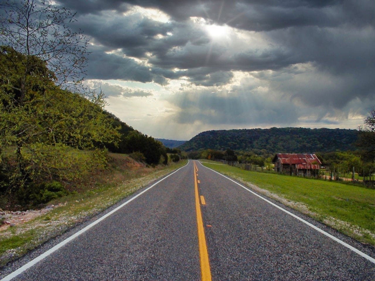 a two-lane highway with a yellow stripe down the middle, trees on the left, a field with an old barn on the right, grey clouds with rays of sunlight piercing thorugh in the sky