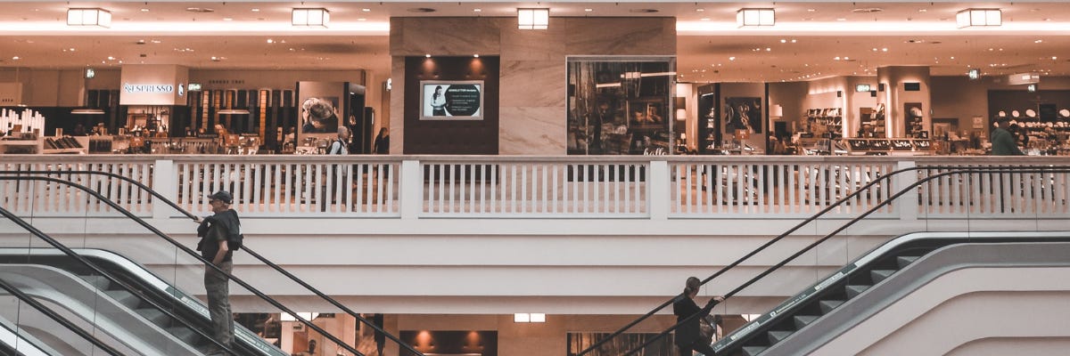 Looking into a large, open store on the second floor of a mall. Two escalators leading up to the second floor are right below it. Two people ride each escalators up.