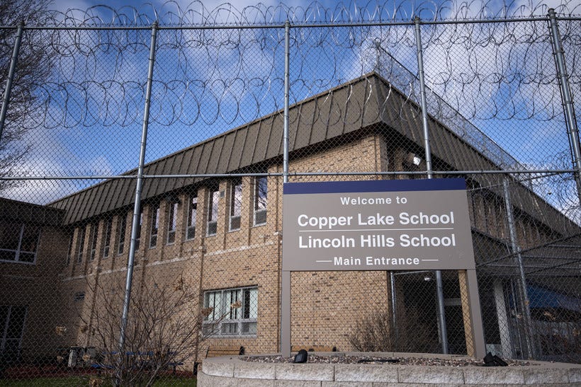 A sign says "Copper Lake School, Lincoln Hills School" in front of a tall fence with barbed wire. 