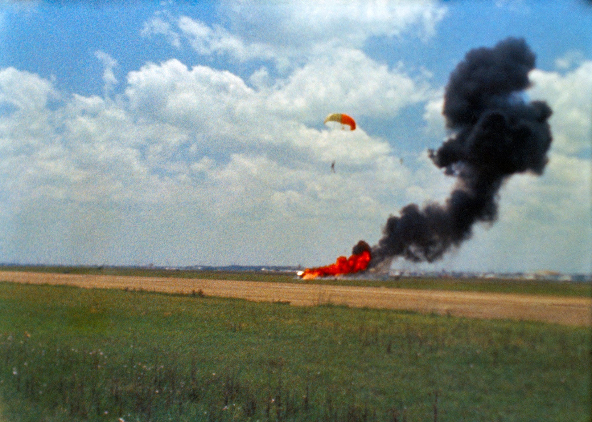 A fiery crash on the ground and a man parachuting above it.