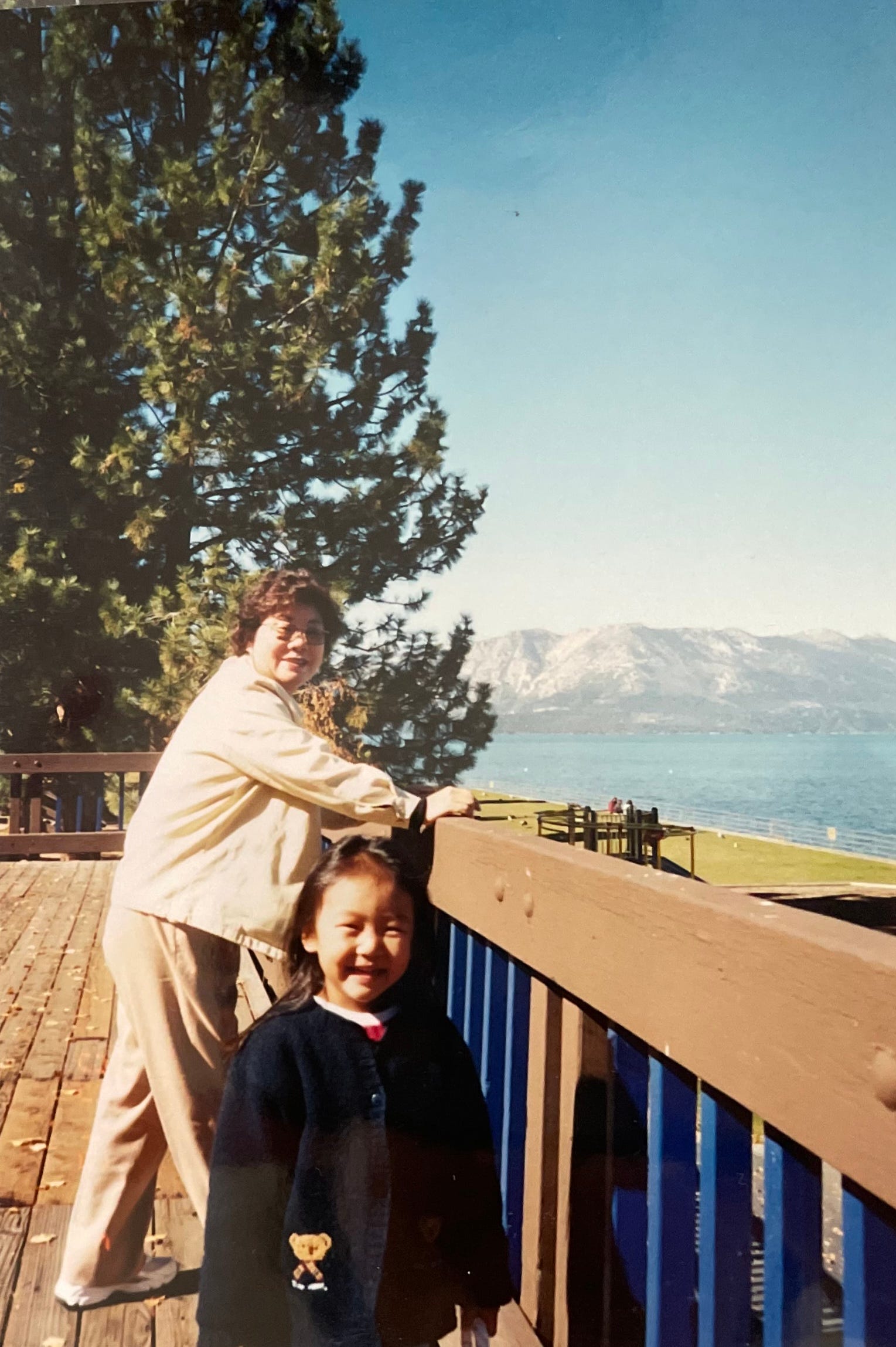 small 4 year old girl (me) wearing a navy cardigan with a small teddy bear embroidred on the pocket. she is in frame from the waist up. her grandma is standing behind her leaning on the wooden balcony of the deck they are standing on. grandma is dressed in head to toe tan jacket and pants, sneakers, and sunglasses. both are smiling at the camera. behind them both is a large pine tree, snowy mountains, and a very blue body of water.