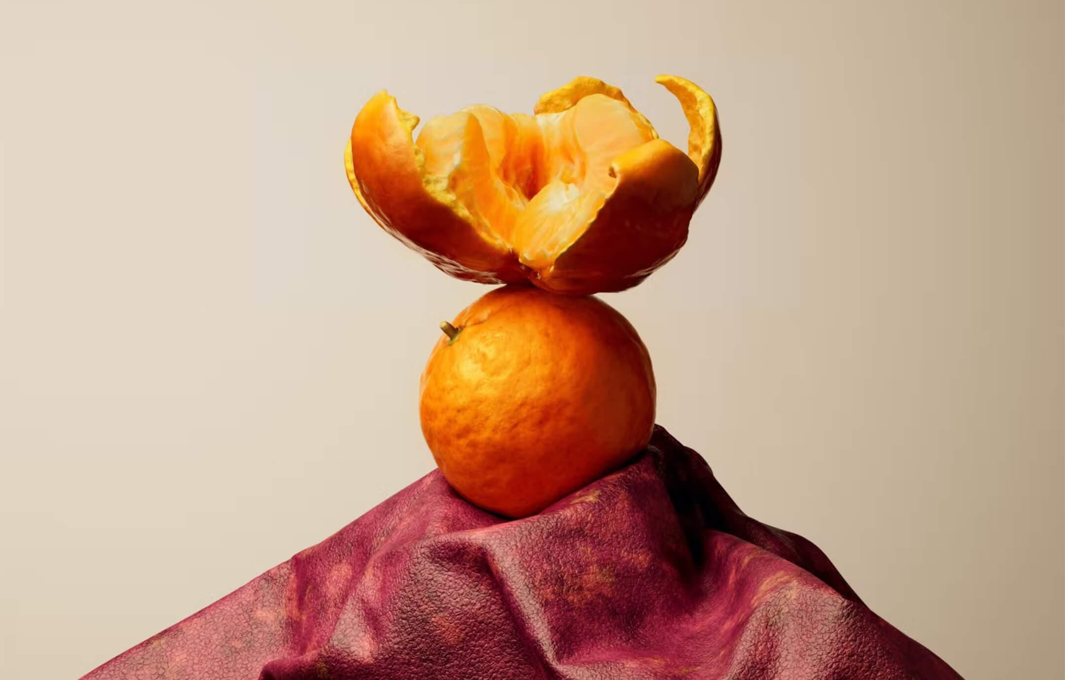 A still-life picture in which an orange rind balances on an orange which balances on a small mount of a dark-red mottled material that looks like leather