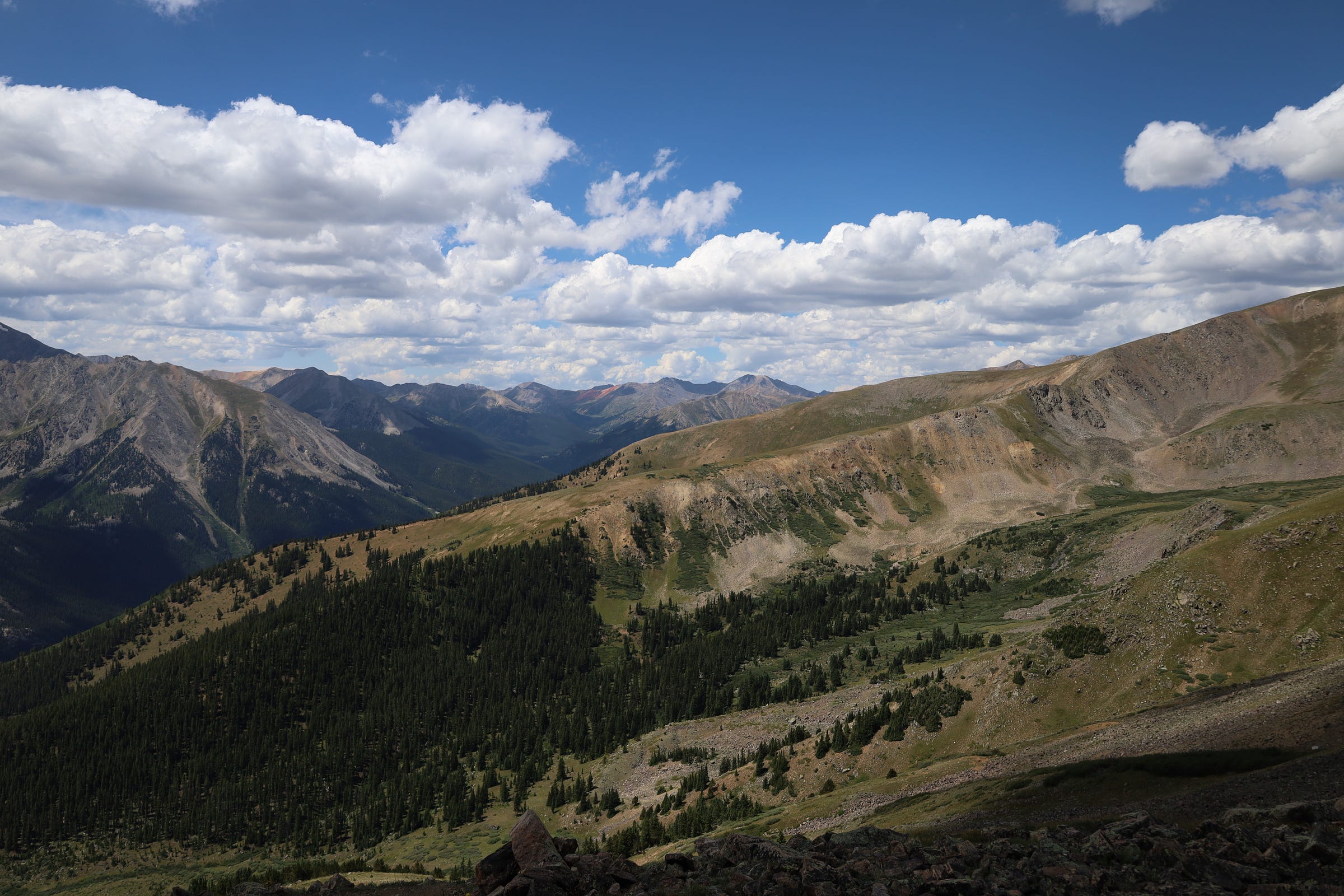 looking back down at the valley, due south of Mt. Elbert. The tree line is far below. puffy white clouds float overhead.