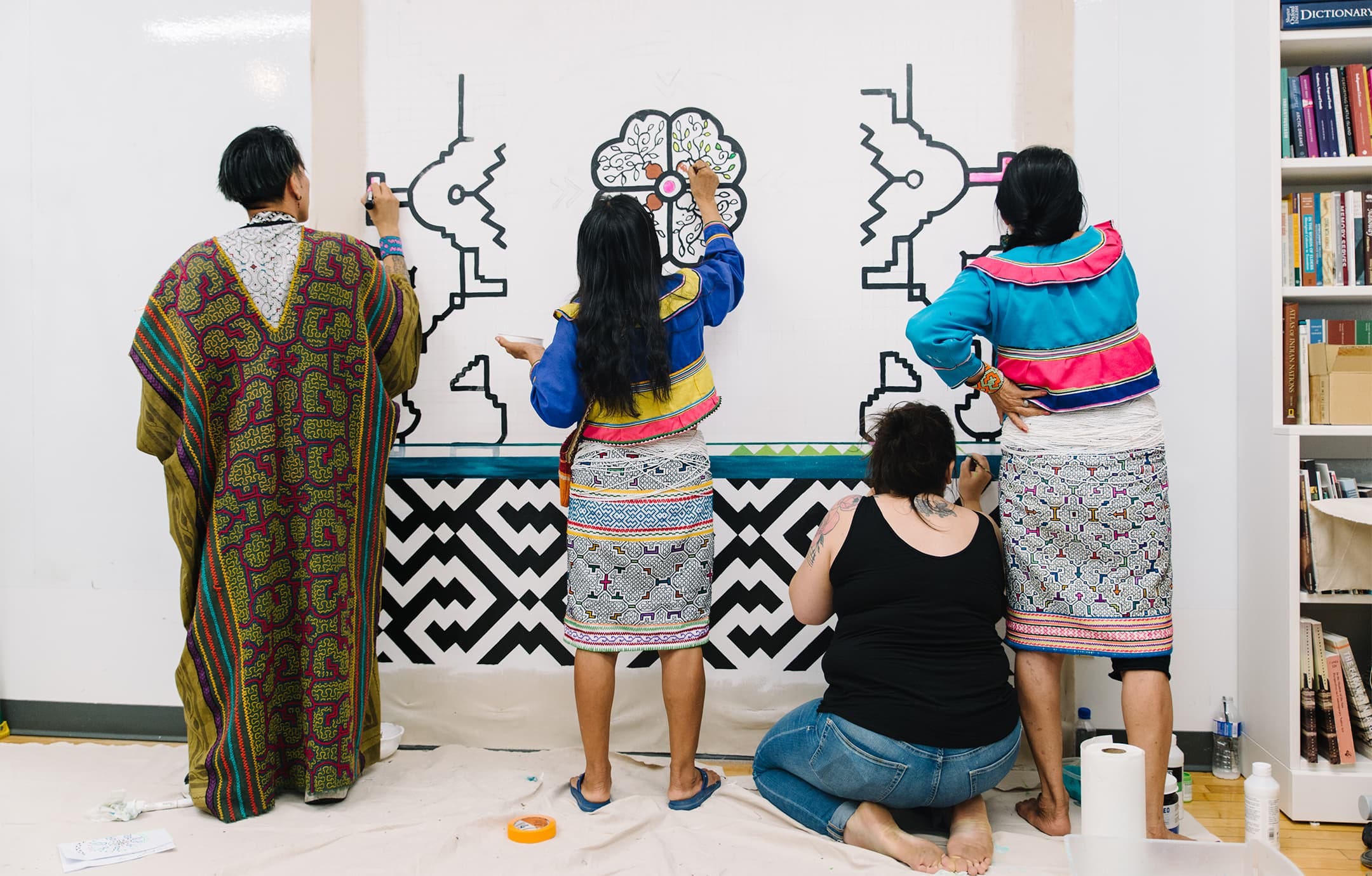 Four figures, three of whom are standing and wearing Indigenous dress and another on their knees in jeans and a black tank top, outline and color in the beginning of a wall design that has distinctly Indigenous graphic markings. (Photo: Stef & Ethan)