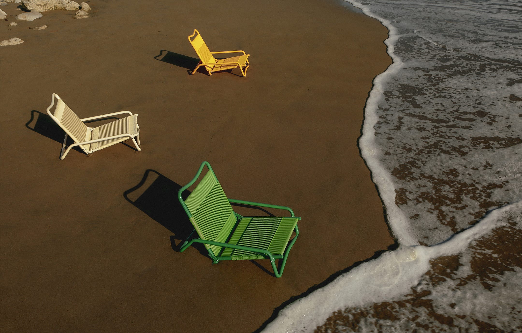Green, cream, and yellow Leisure Chairs by ITA Leisure sitting in the sand as a wave laps the beach