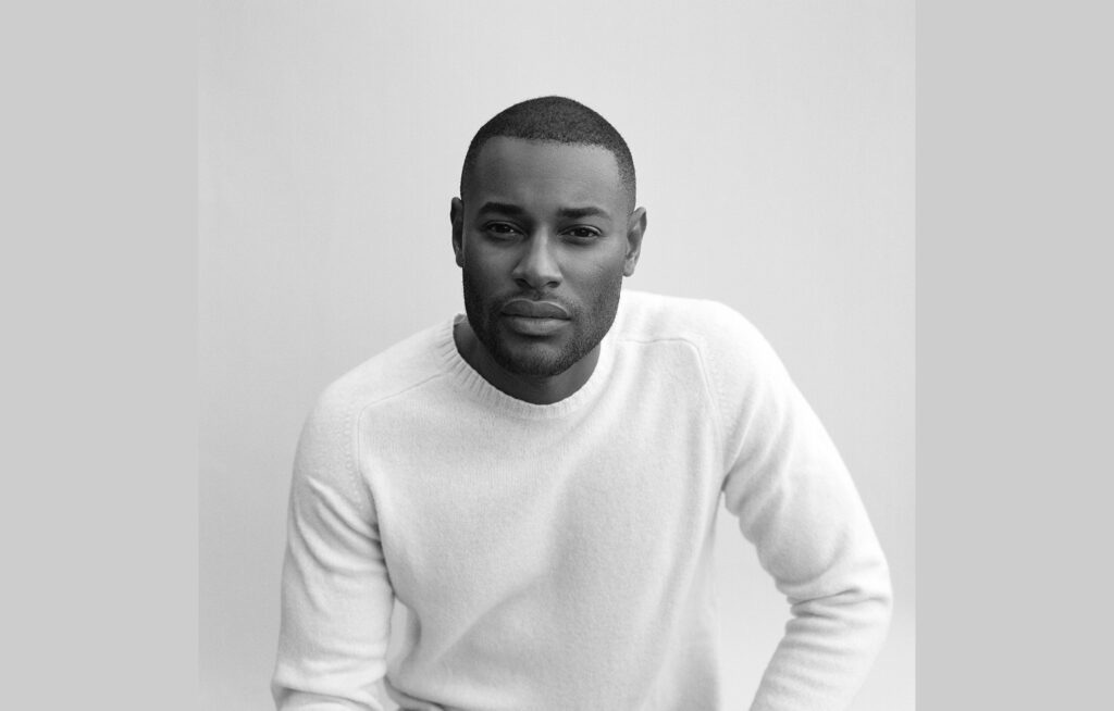 Photograph of model-turned-entrepreneur Roger Dupé. A Black man with close-cropped hair and stubble on his face, he peers into the camera while wearing a white sweater.