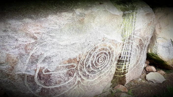 Greyish oblong kerbstone at Knowth, 3 etched designs, one which looks like a letter E, followed by circular swirling motif, and another which is hard to describe but reminds me of a snail.