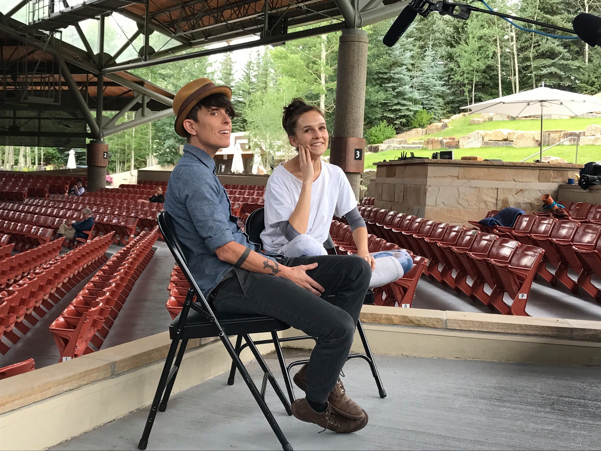 Andrea Gibson and Lauren Lovette sit on an outdoor stage at the Vail Dance Festival smiling at the camera, the chairs around them are empty and you can see mountains in the background.