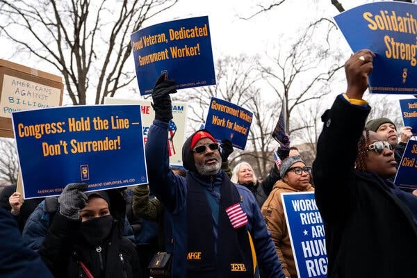 People dressed in winter clothing hold up signs with slogans including “Congress: Hold the line — don’t surrender!” and “Proud veteran, dedicated government worker!”