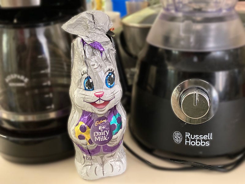 A white-silvery, modest sized Easter bunny made by Cadbury, minus its ears. In the background are kitchen appliances including a coffee perculator and blender.