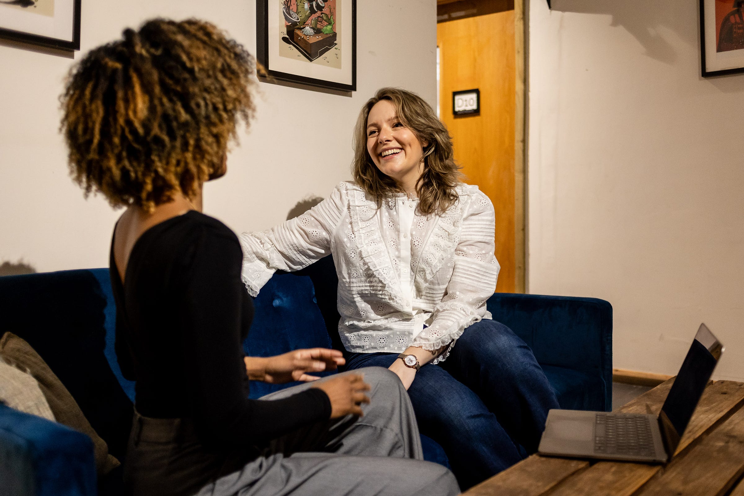 Image description: Ettie’s a white person with light brown hair, sat on a blue sofa indoors. She’s facing the camera and laughing, as she talks to someone who’s facing away from the camera, who has a light brown Afro, and is wearing a black top, grey trousers. 