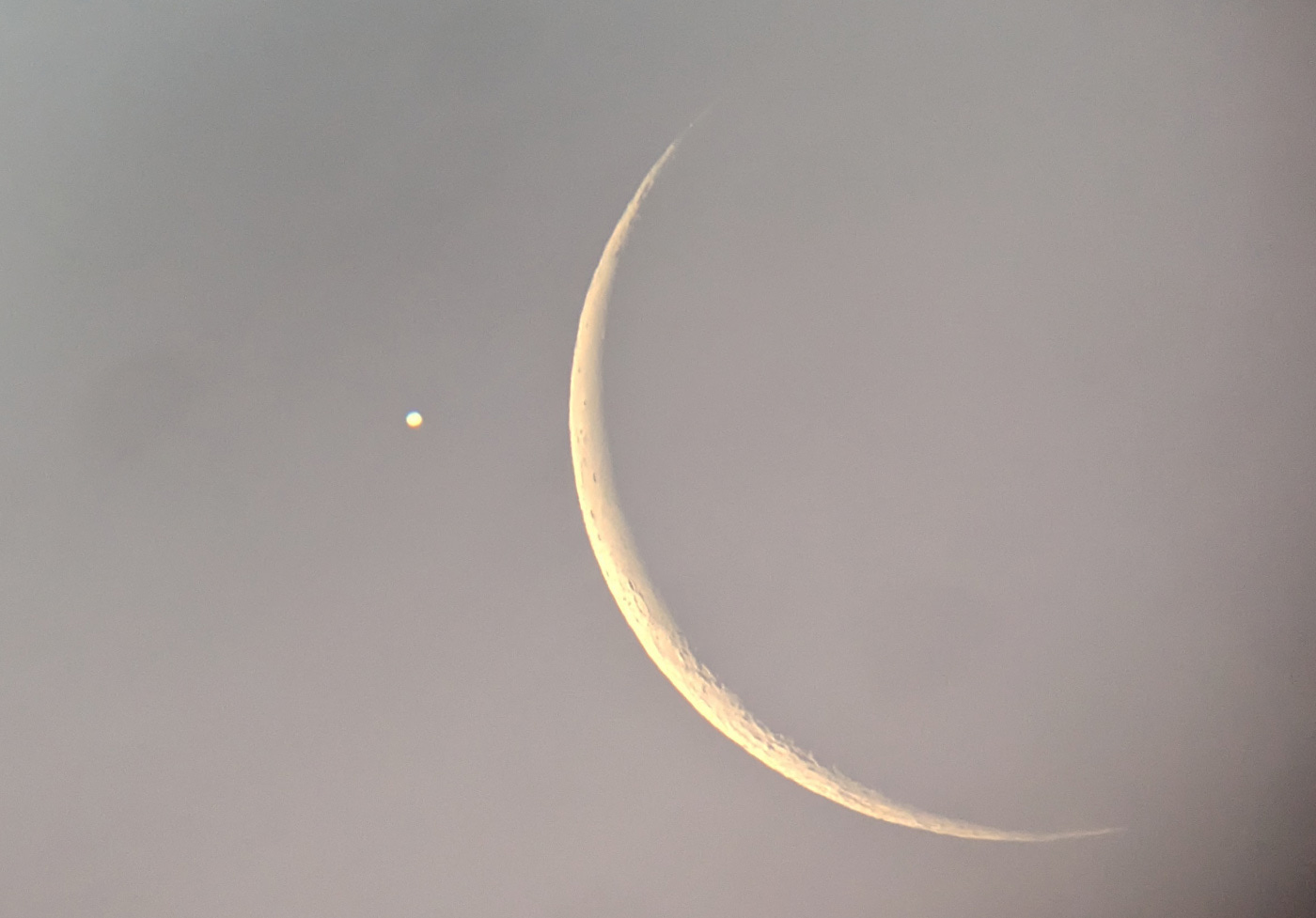 Jupiter appears as a small bright white disk next to the much larger but very thin crescent Moon, both against a gray sky at dawn. A few lunar craters are visible. Jupiter appears as a small bright white disk next to the much larger but very thin crescent Moon, both against a gray sky at dawn. A few lunar craters are visible.