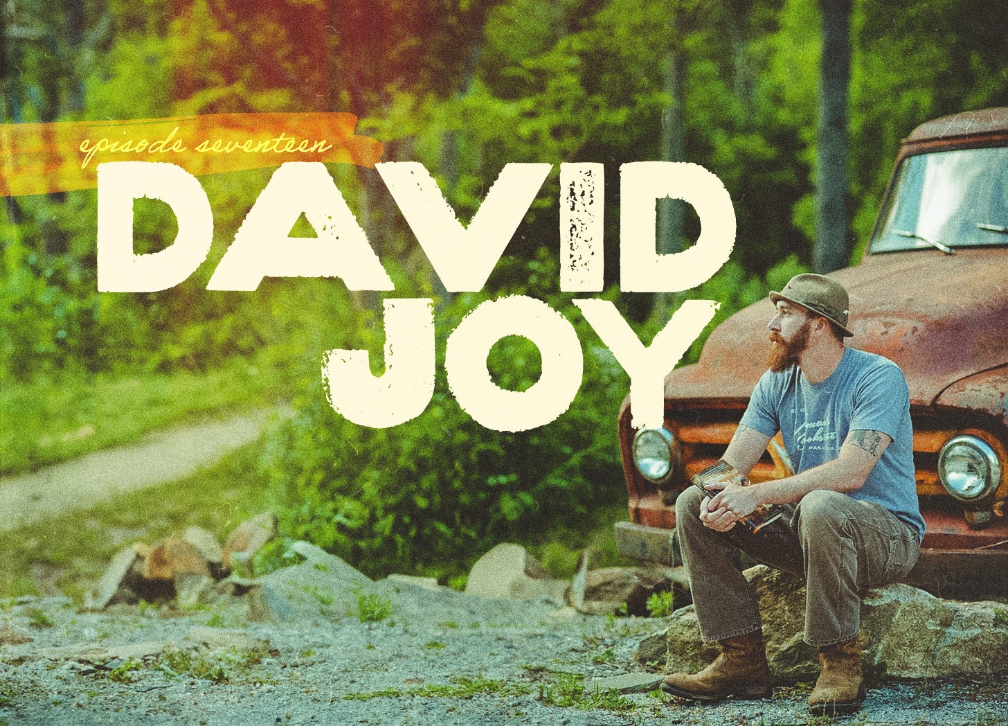 A man holds a book while sitting on a large rock in front of an old truck in the mountains.