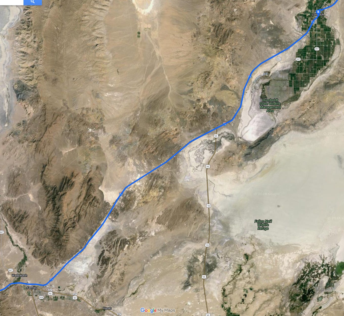 Satellite view of the Nevada desert from Fernley in the lower left to Lovelock in the upper right from Google Maps. A blue line representing the route runs from the lower left to upper right of the image. There are a couple of roads running north and south into Fernley and Wadsworth in the lower left. There is another road - labeled 95- running up from the bottom center to the blue line. There are no other roads. The majority of the image is barren desert - white sands, tan soils, a dark ridge of hills in the lower left, and a line of green near Wadworth in the lower left marking a river. In the upper right, leading to Lovelock, is an oblong area - a green grid of irrigated fields. The blue line extends beyond Lovelock orr the upper right corner of the image.