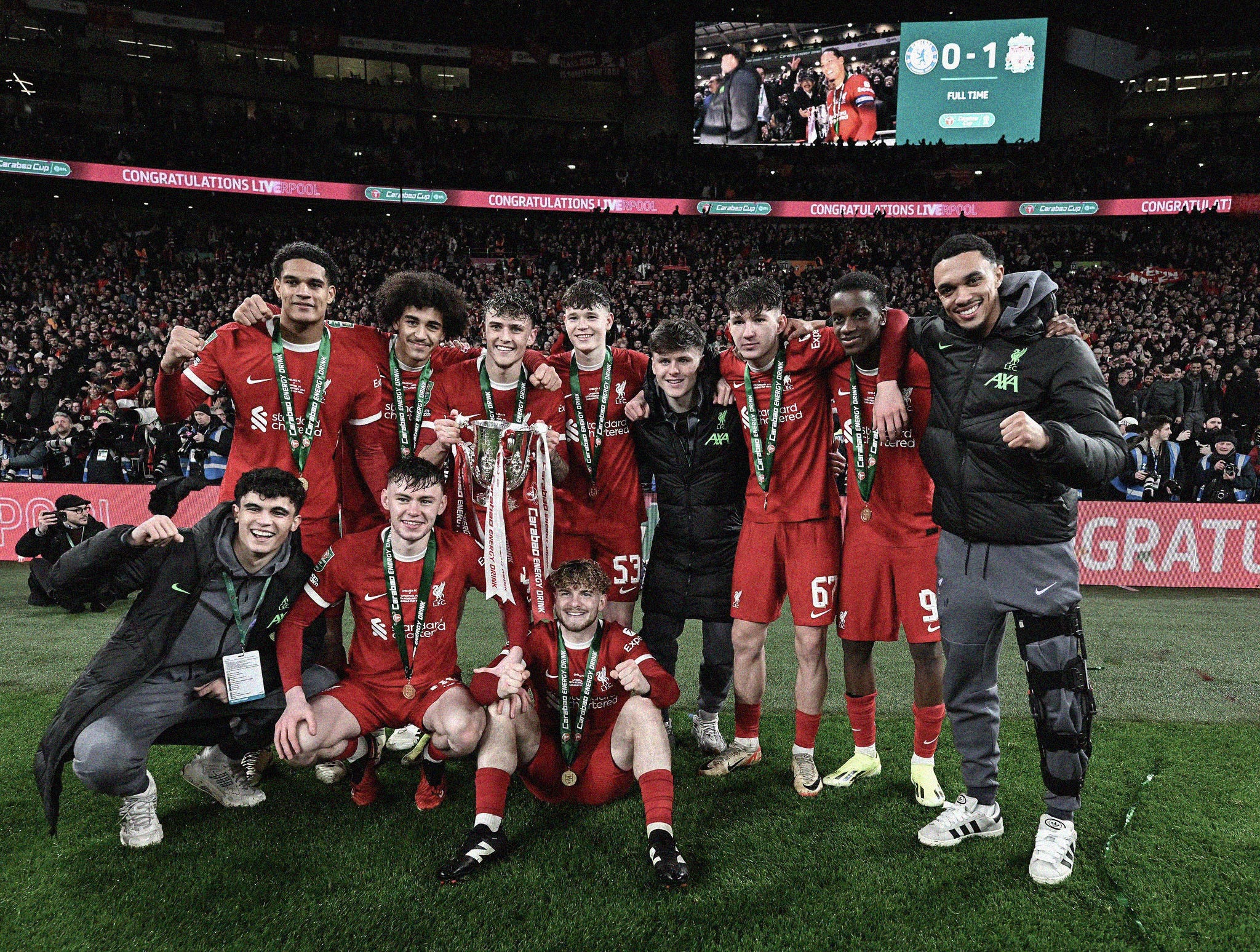 A photo of Liverpool's academy players with the League Cup trophy. They're wearing an all-red kit, stood arm-in-arm, celebrating in front of the Liverpool end at Wembley.