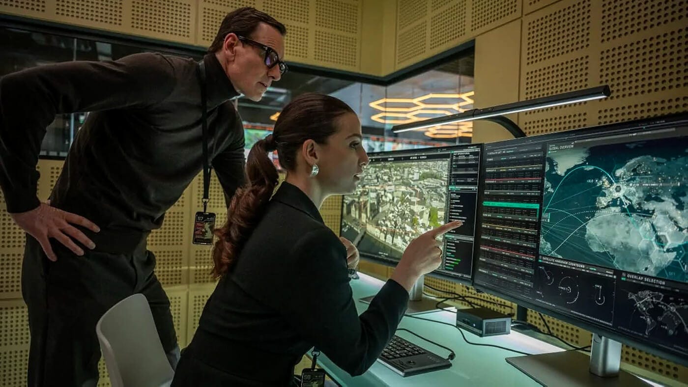 A man and woman in dark clothing study a bank of monitors displaying satellite surveillance data and global heat maps. The woman points at the screen as they hover over the console in a modern glass-walled room.