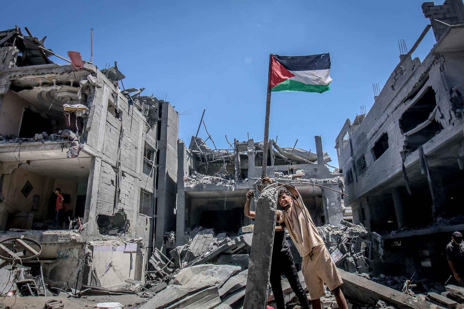 Palestinian youths raise the Palestinian flag amidst the rubble of their homes, destroyed by an Israeli airstrike on a residential block in Al-Bureij Camp. Gaza Strip, 2024. Photo by Saeed Mohammed Jaras