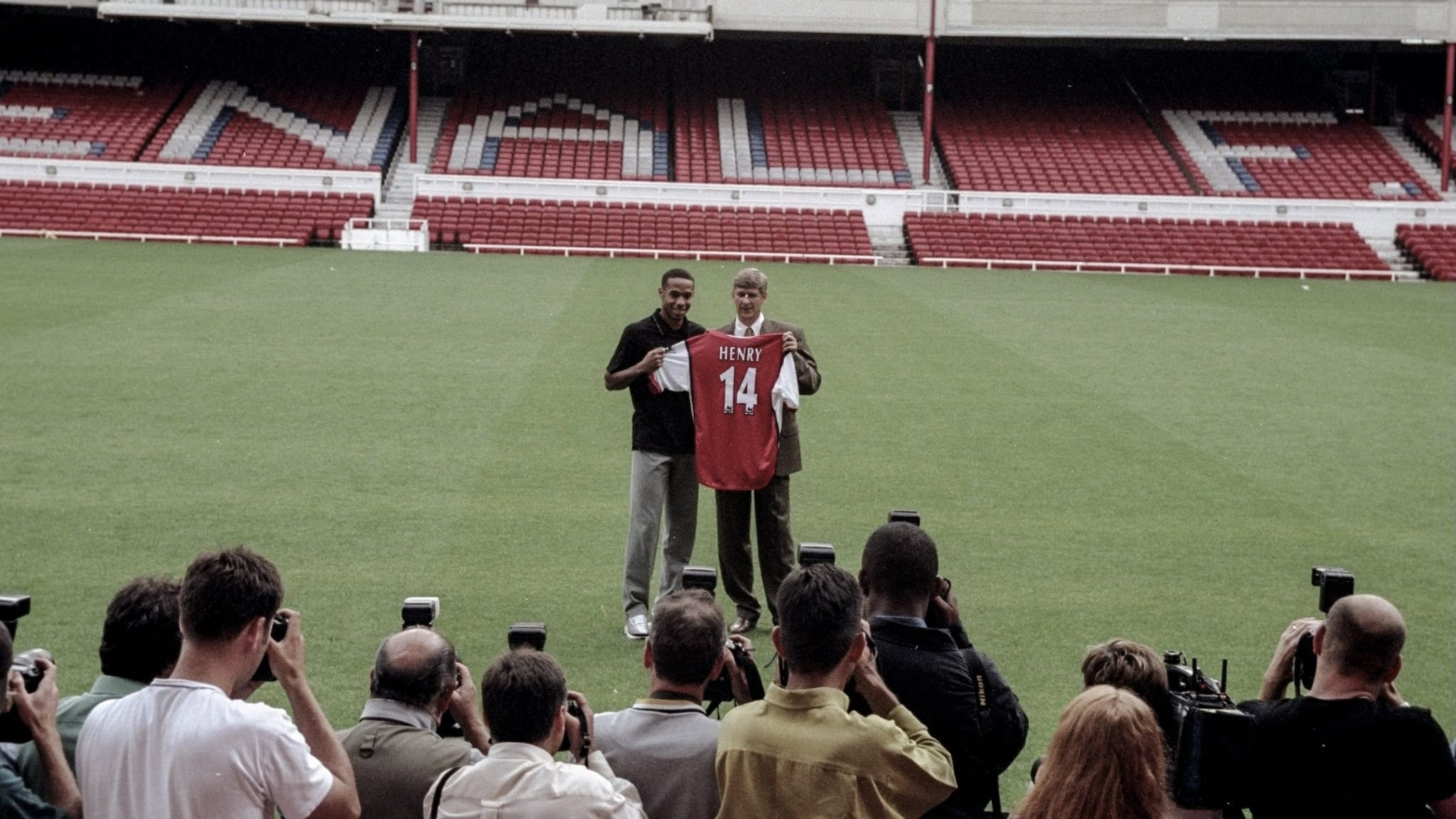 A wide-frame photo of Thierry Henry and Arsène Wenger holding up an Arsenal shirt, stood on the Highbury pich, with a line of photographers in the foreground