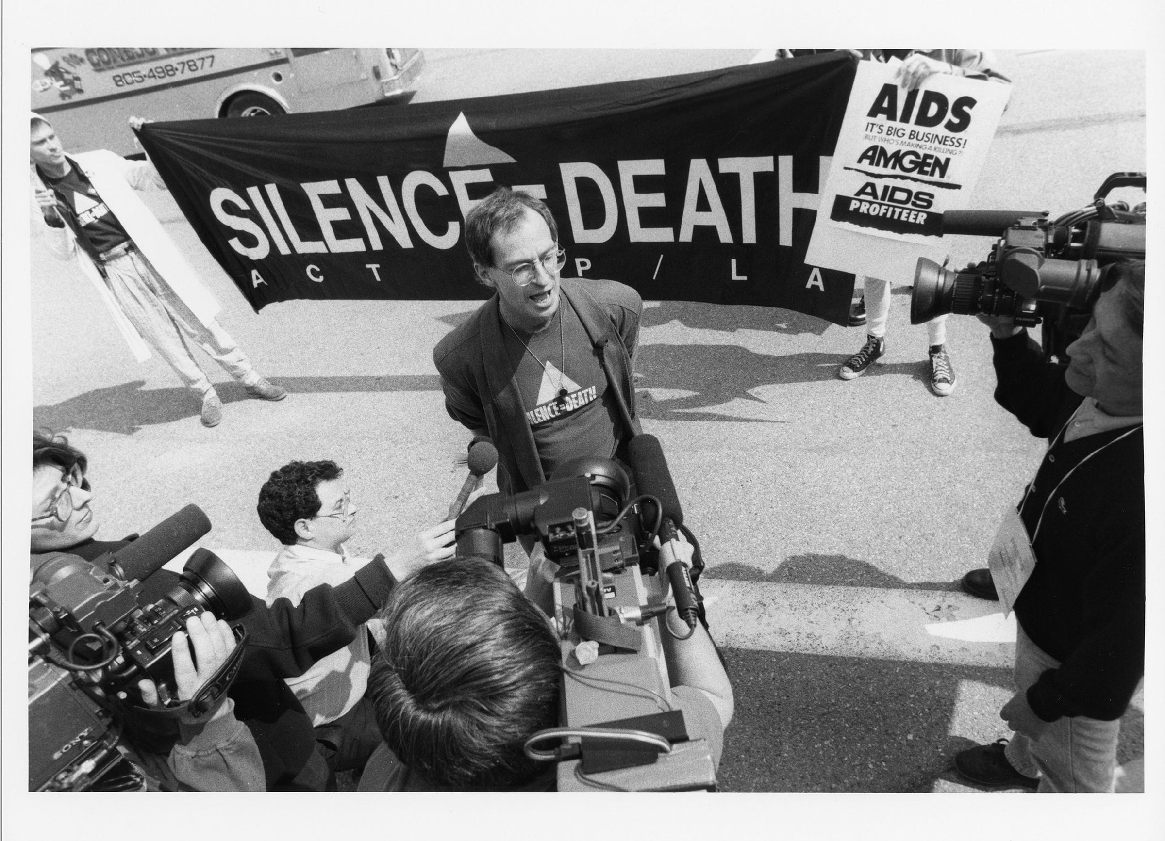 A black and white photo of David Lacaillade wearing a Silence = Death T-shirt while surrounded by members of the press with cameras and microphones. Two ACT UP activists stand behind him holding a “Silence = Death” banner and a sign reading “AIDS: It’s Big Business! But Who’s Making a Killing? AMGEN, AIDS Profiteer.”