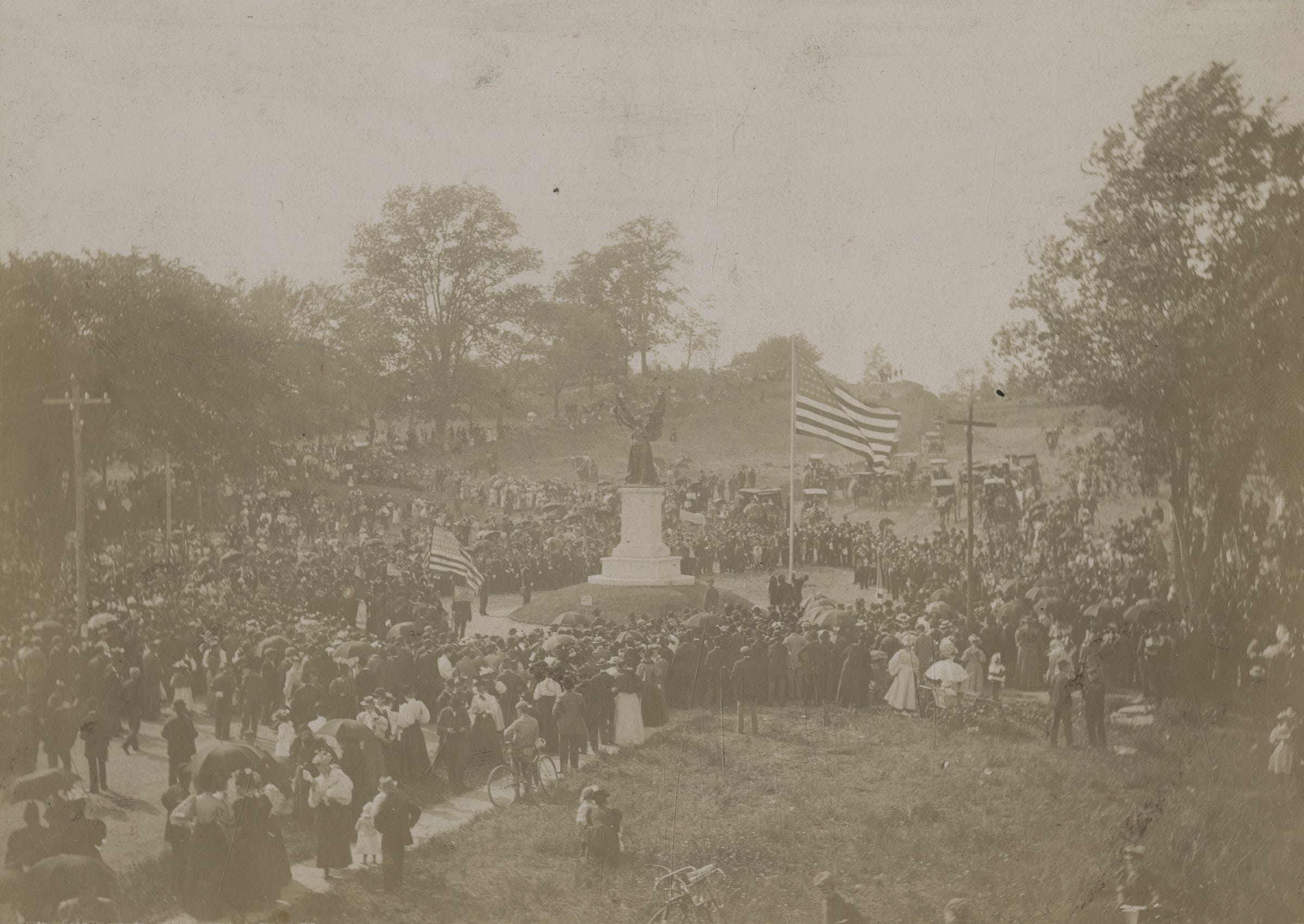 Photograph shows a crowd gathered around the Jamaica Soldiers' and Sailors' Monument at the dedication ceremony. There are trees and hills and many people, horse carriages, flags blowing in the wind. That only a few people have umbrellas suggest it was a warm sunny day.