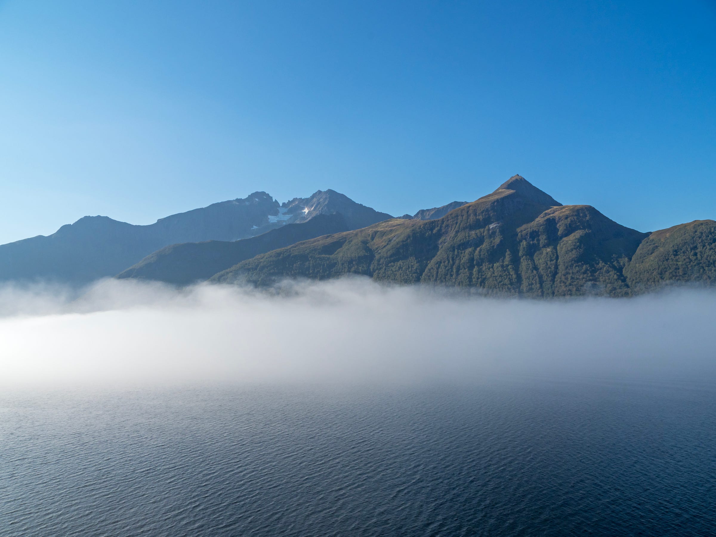 Original photographs of mountain peaks with a small area of snow rising above a Norwegian fjord.  There is a thick layer of white mist above the water of the fjord which is obscuring the lower part of the mountains.  There is a clear dark blue sky above.