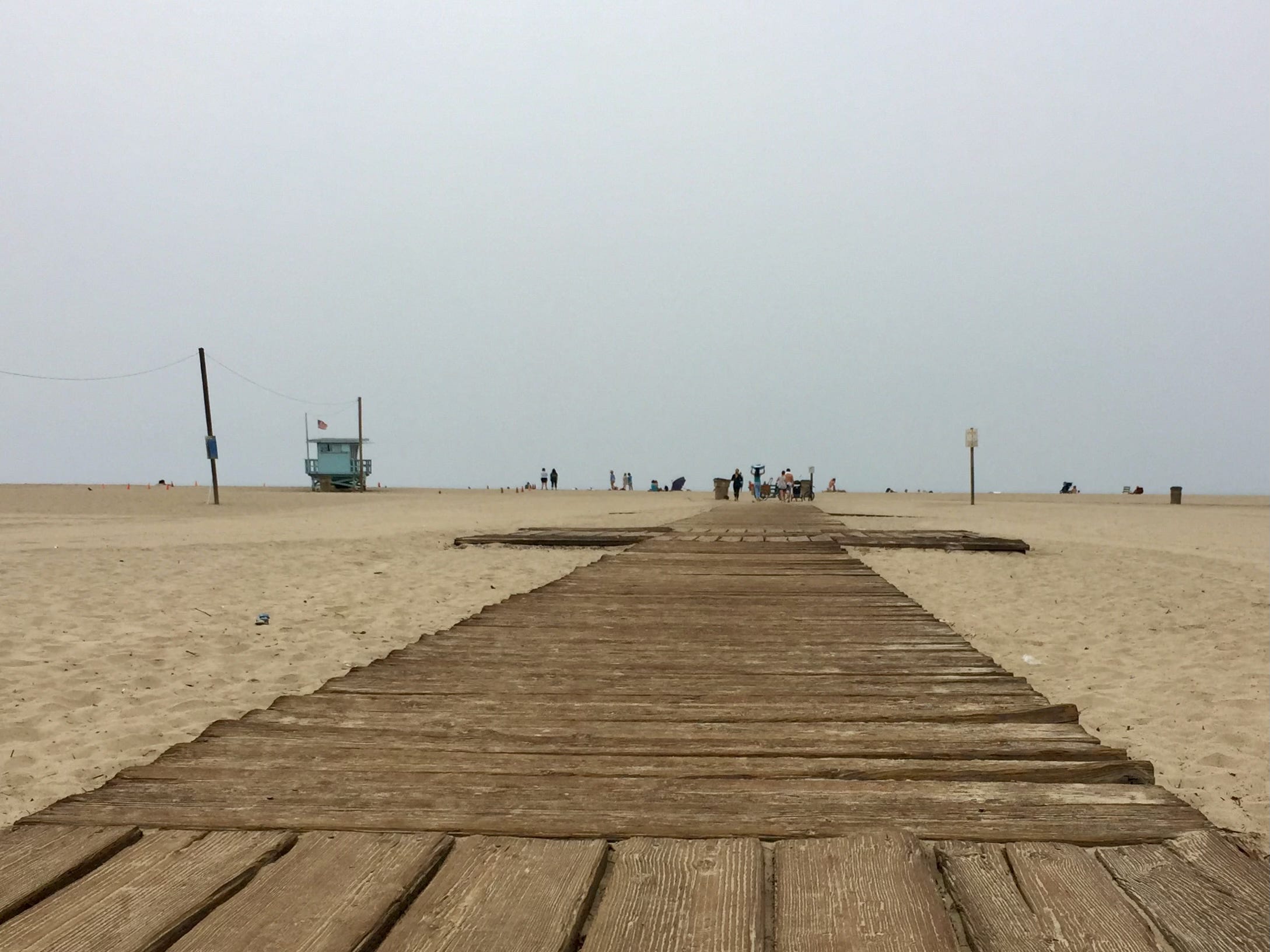 A boardwalk reaching across Santa Monica beach on an overcast day. A blue lifeguard stand in the distance. 