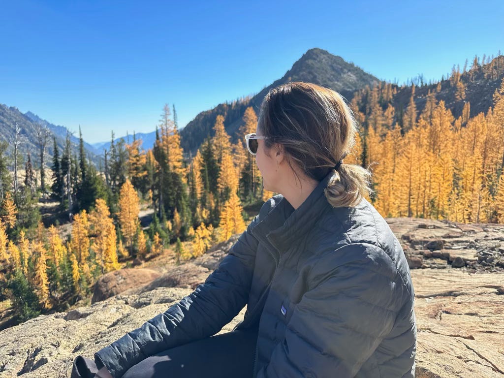 A woman sits on a rock and looks over her right shoulder to a forest of yellow larches behind her in the distance against a blue sky.