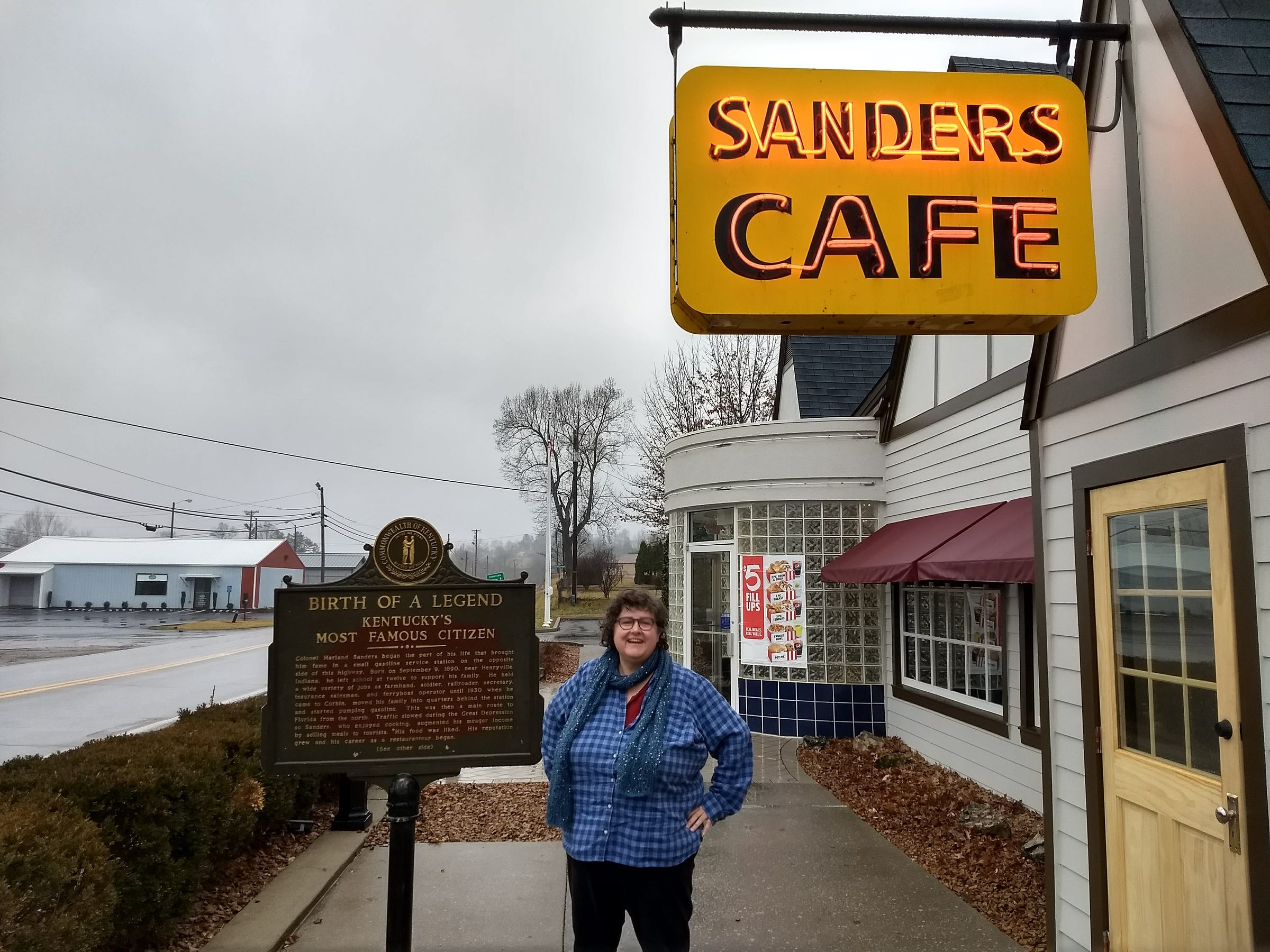 Annette standing next to historical info panel in front of buiilding with Sanders Cafe neon sign, on rainy day