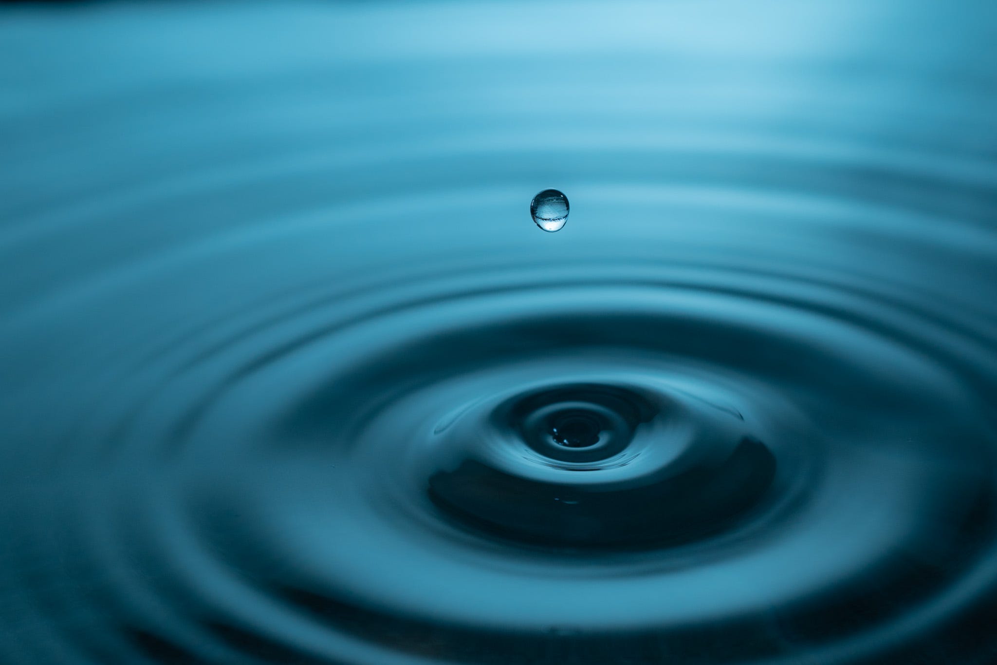 A water drop splashes into a pool of water creating a ripple effect with a reflection of the water in the droplet. A light filter causes water and droplet to look blue.