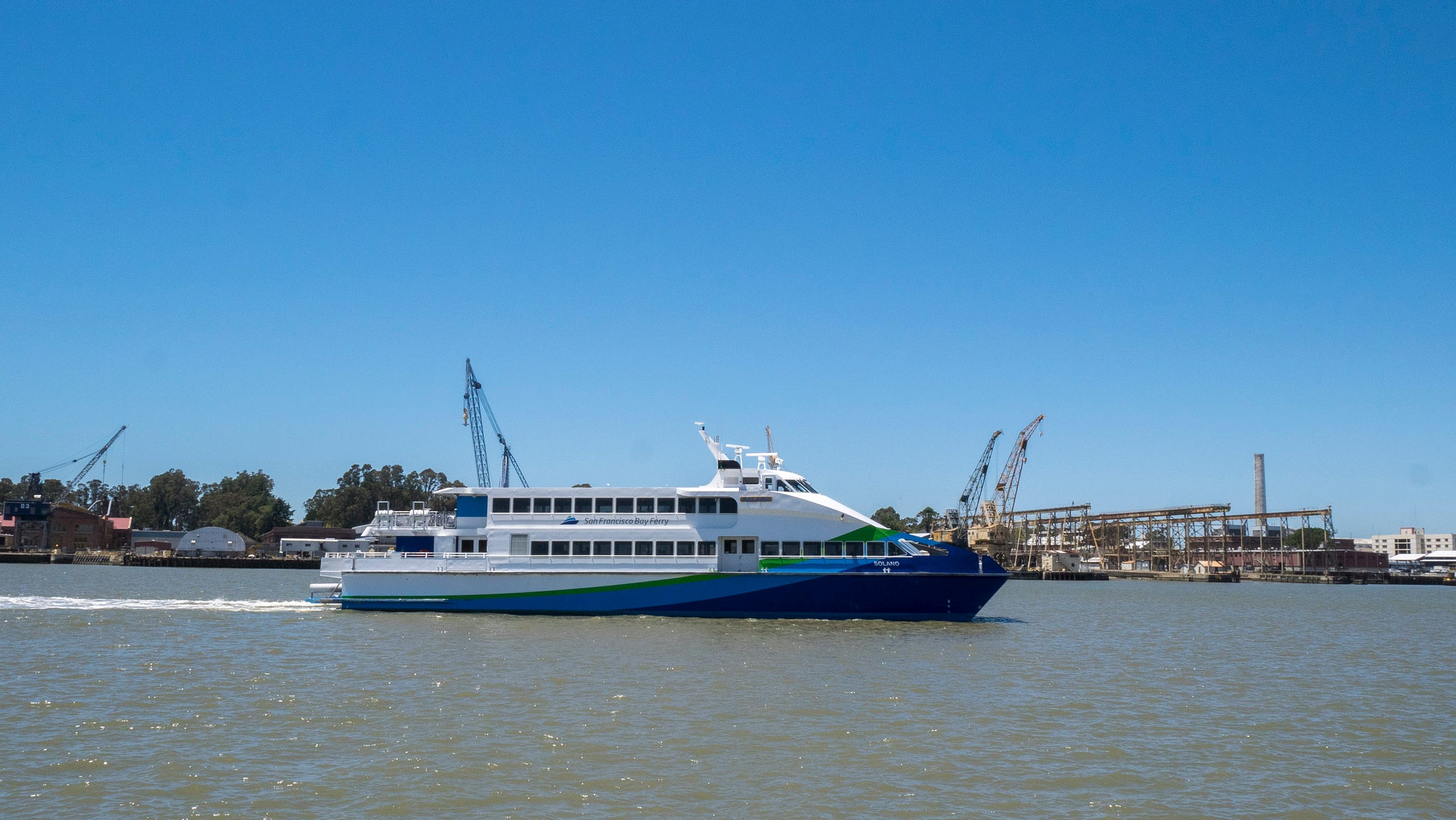 A double-deck modern passenger ferry (white with blue and green streaks rising from the tail to the bow) pulls into the ferry terminal at Vallejo, California. Shipping cranes rise in the background.