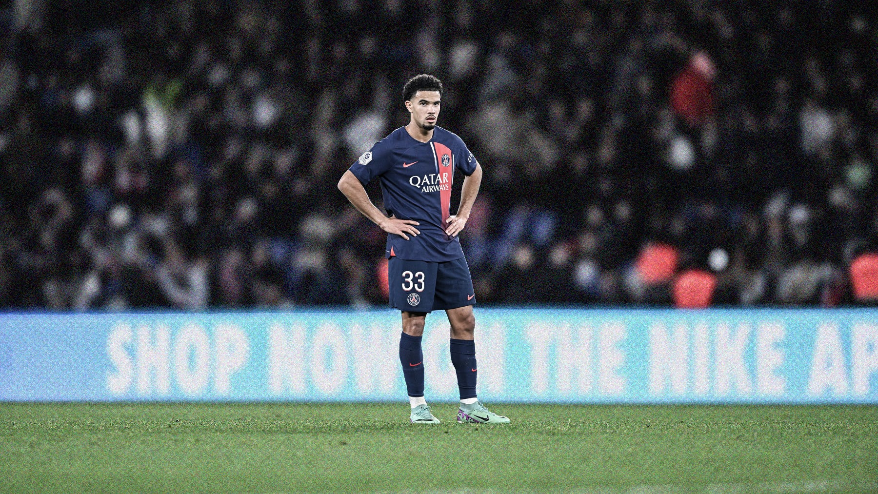 A wide-frame photo of Warren Zaïre-Emery in a navy Paris Saint-Germain kit, stood with hands on hips in front of a blurred crowd background