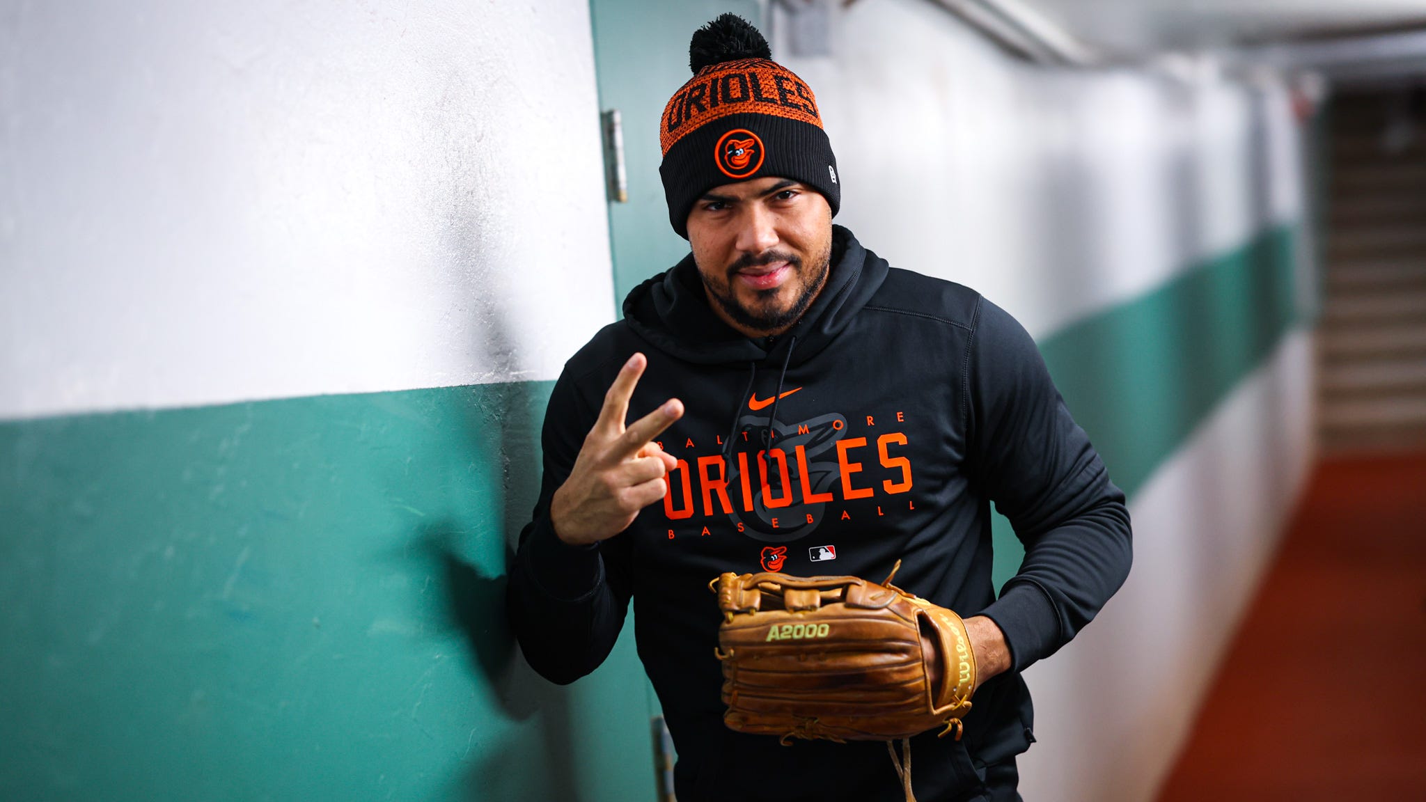 Anthony Santander holds up a peace sign while walking in the tunnel to the field. He's wearing an Orioles beanie and sweatshirt. 