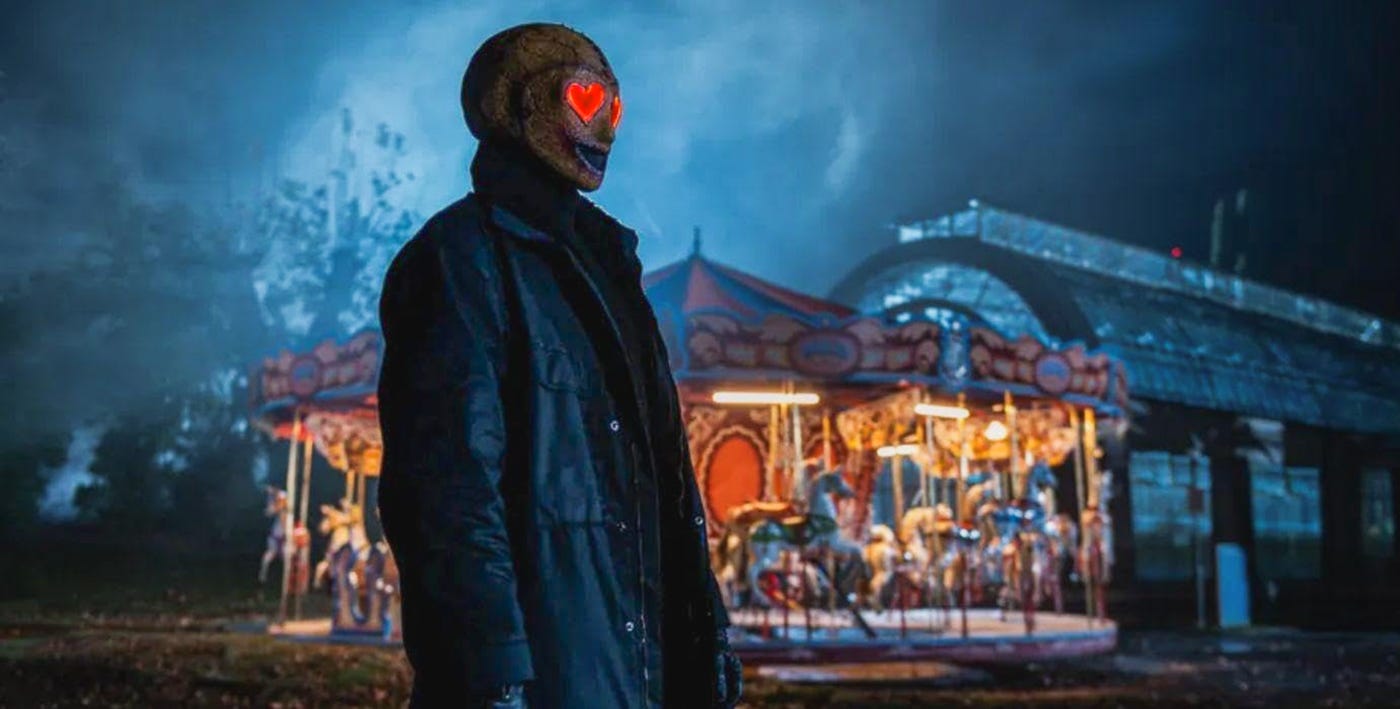 A masked figure with glowing red heart-shaped eyes stands in front of a carousel at night. A masked figure with glowing red heart-shaped eyes stands in front of a carousel at night.