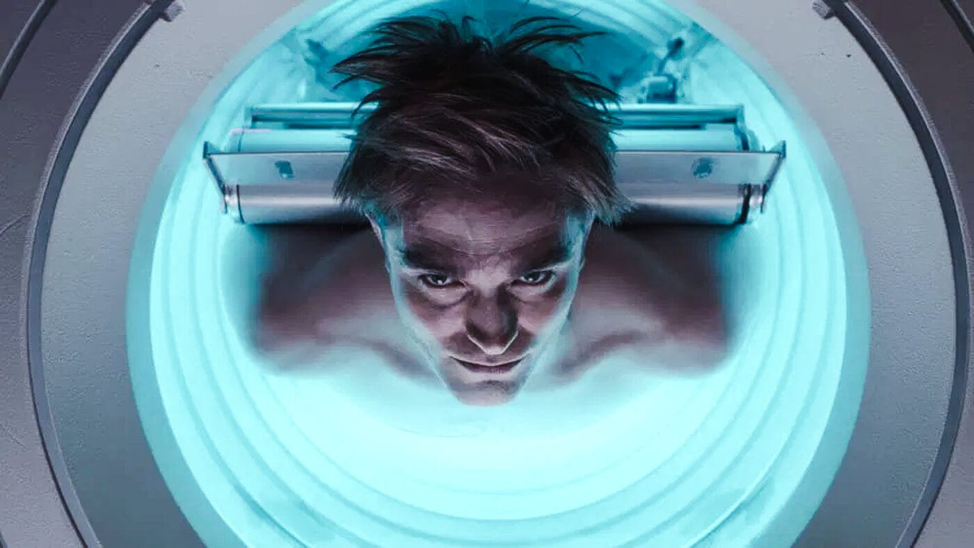 Overhead shot of Robert Pattinson lying naked in a glowing blue cloning chamber, eyes open and staring upward as he reawakens inside a circular pod.