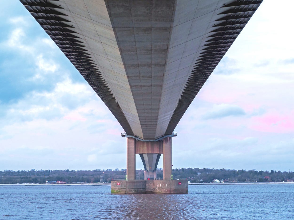 Photograph taken directly below the Humber Bridge, showing the underside of the bridge stretching away into the distance above the estuary. You can see through one of the support towers. There is a blue and pink cloudy sky above just before sunset.