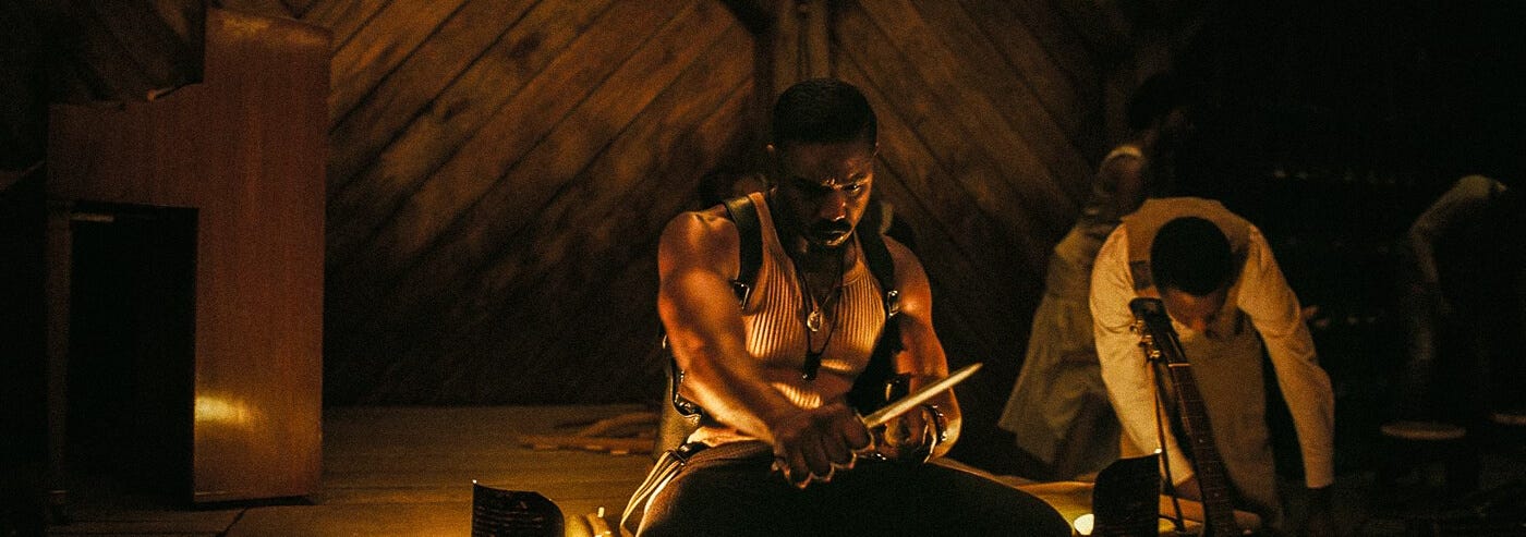 Michael B. Jordan sits while sharpening a stake, bathed in warm stage lights, flanked by musical instruments and fellow townsfolk—tension hangs heavy in the air.