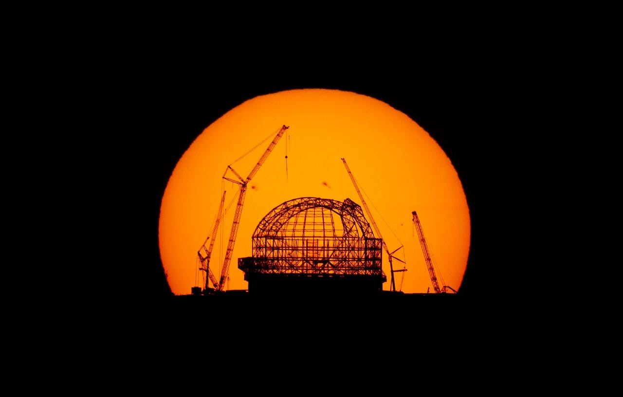 The Sun appears as a swollen orange disk behind the partially completed scaffolding of the observatory dome, with huge cranes silhouetted on either side of it. The Sun appears as a swollen orange disk behind the partially completed scaffolding of the observatory dome, with huge cranes silhouetted on either side of it.
