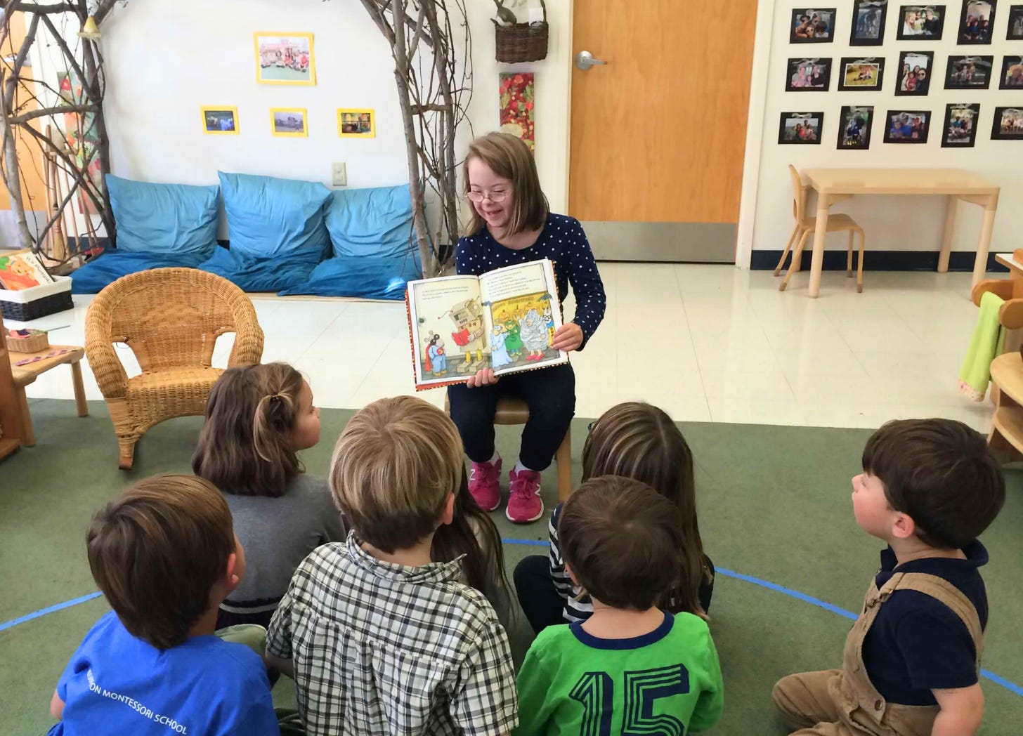 Penny in 2015 sitting in a small chair in a classroom and reading a book to students.