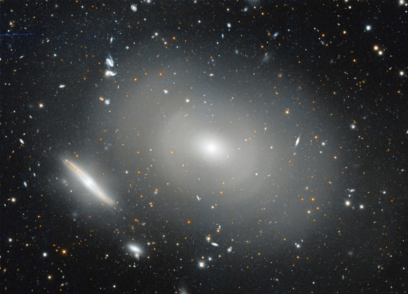 An image showing a close-up of one elliptical galaxy that looks like a cotton ball, surrounded by several concentric rings of light. Next to it is an edge-on disk galaxy, and there are dozens of background galaxies visible as well. An image showing a close-up of one elliptical galaxy that looks like a cotton ball, surrounded by several concentric rings of light. Next to it is an edge-on disk galaxy, and there are dozens of background galaxies visible as well.