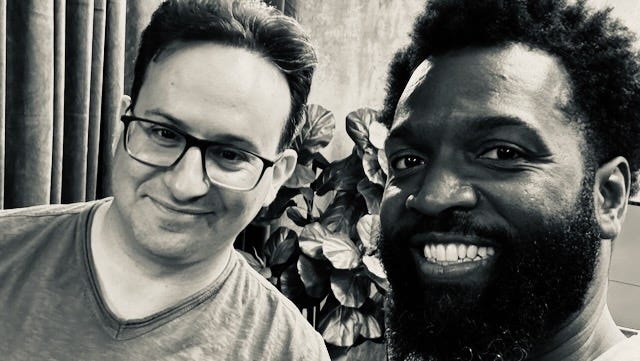 Photo of Jared, left, and Baratunde, right. Filter on photo of Silvertone black and white. Jared has glasses and a t-shirt. Baratunde also has a t-shirt. The plant in the photo is fake. 