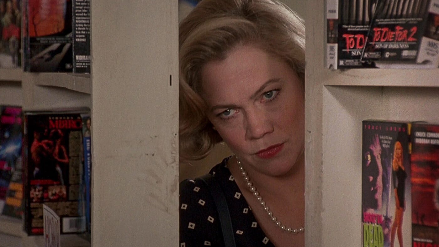 Kathleen Turner peeking menacingly from a video store back room, framed by VHS tapes and seething judgment. Kathleen Turner peeking menacingly from a video store back room, framed by VHS tapes and seething judgment.