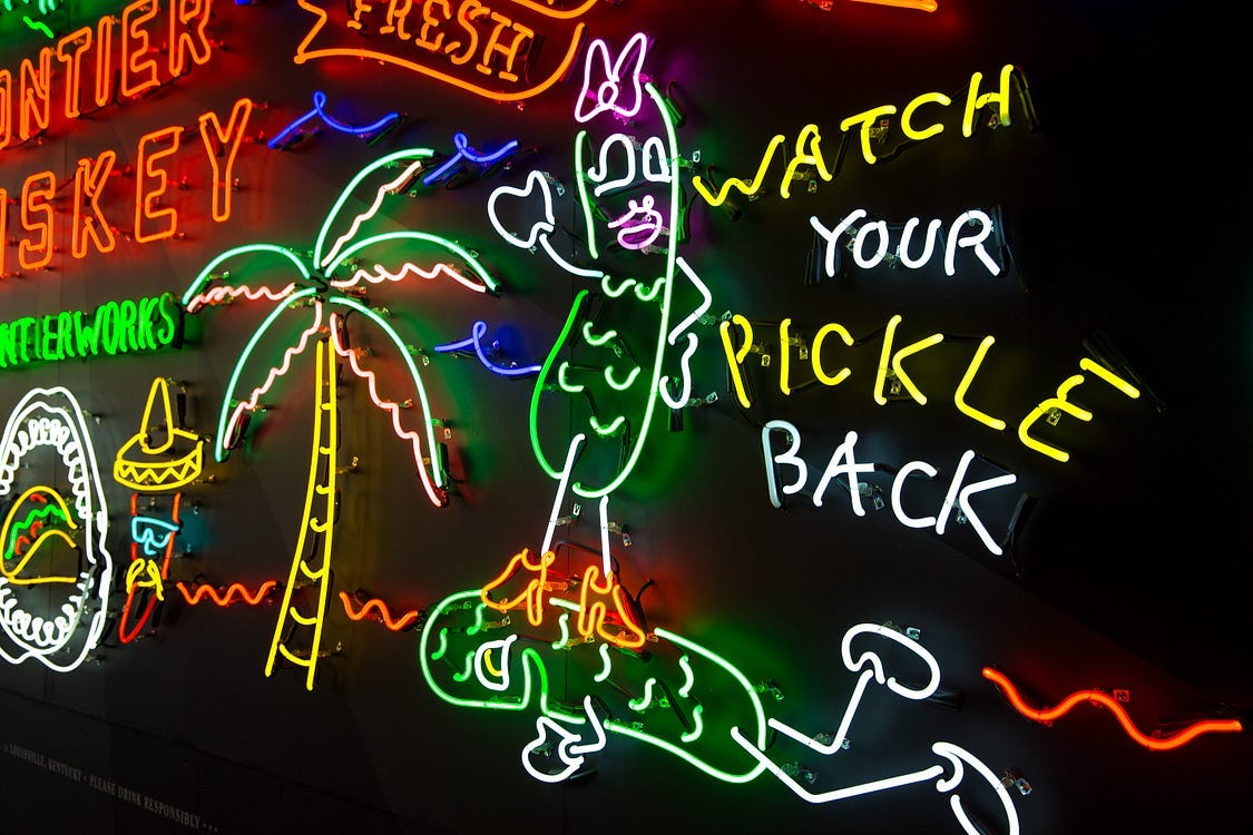 A neon sign depicting a female pickle standing on the back of a male pickle. reads: Watch your pickle back. It’s a whiskey advert or bar sign.