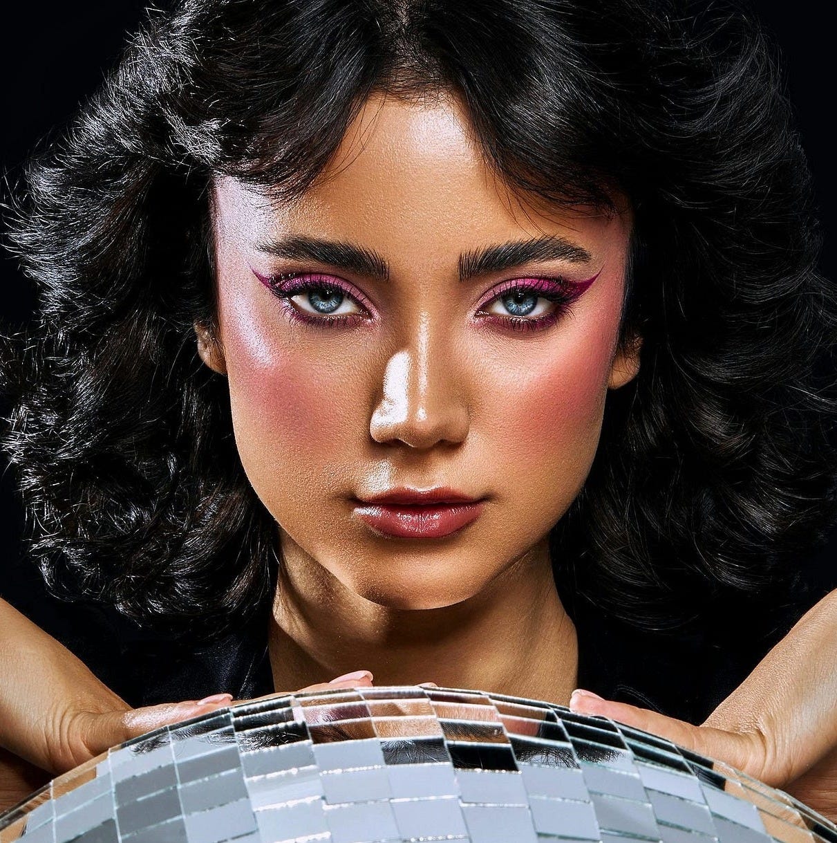 A front-on image of a woman with black hair, blue eyes, and pink eyeshadow/eyeliner. She’s posing with her hands pressed on a disco ball, the backdrop for the image is black. A front-on image of a woman with black hair, blue eyes, and pink eyeshadow/eyeliner. She’s posing with her hands pressed on a disco ball, the backdrop for the image is black.